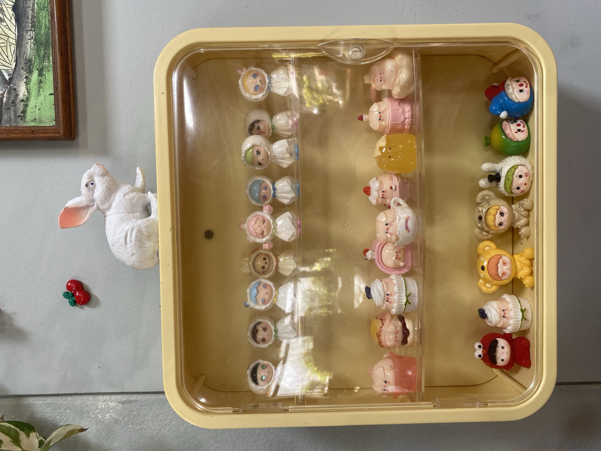 A top-down shot reveals a collection of small, whimsical figurines contained within a yellow plastic box, situated indoors. The box is divided into three sections, each housing a different type of figurine. The first section contains figures with white bonnets and various colored hair. The second section features figurines resembling desserts or sweets. The third section includes characters such as bears, cupcakes, and other whimsical creations.
Adjacent to the box on the left is a white rabbit figurine and a small red cherry decoration. To the top left of the image, a framed picture hangs on the wall. Plant foliage is visible in the lower left corner. The scene is clean, and the light is even.