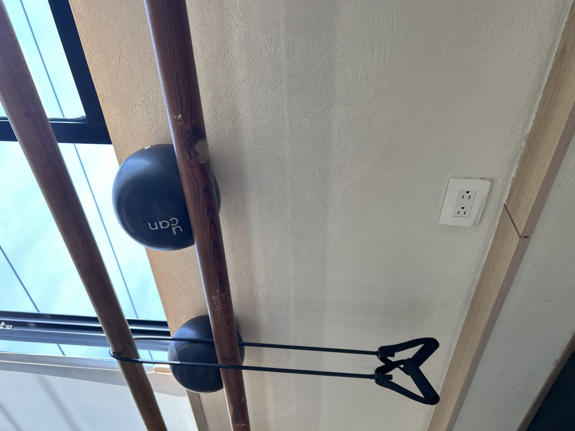 The image shows a view looking upwards, possibly inside a building. Two dark-colored exercise balls are wedged between wooden bars. The upper ball has the letters "ucan" printed on it. A black resistance band with handles is stretched between the lower ball and the upper right corner of the frame. In the background, there's a window with visible daylight. An electrical outlet is present on the wall in the upper right portion of the image.