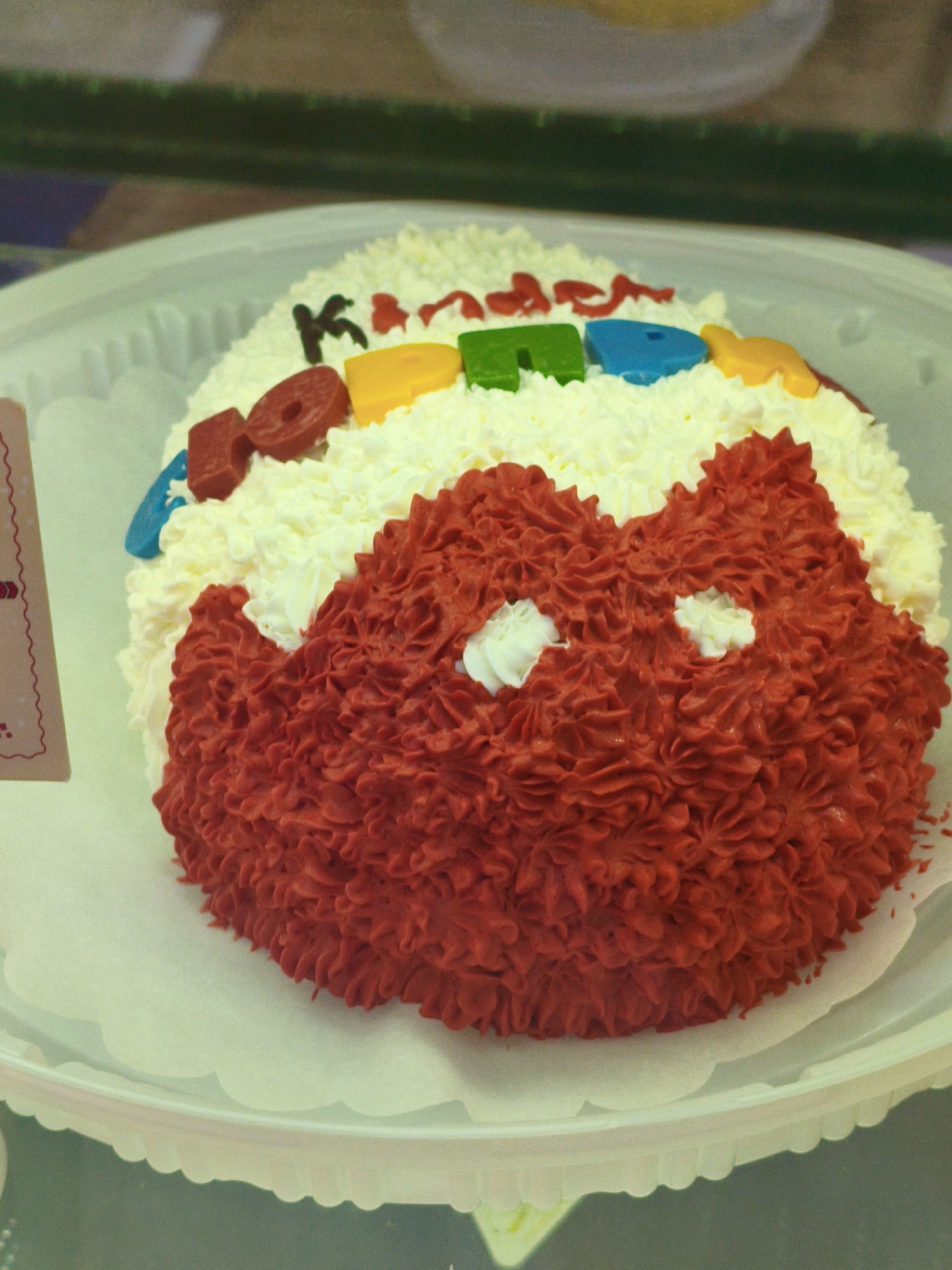 The image shows a close-up view of a red and white cake decorated with candy letters and shapes. The cake is round and covered in white frosting, with a section of red frosting forming a textured pattern on the front. Colorful candy letters spelling out "KARA" are placed on top of the white frosting, along with a blue, green, and yellow candy piece. The cake sits on a white scalloped plate, which is resting on a surface that appears to be a display case. The background is blurred, with hints of greenery and other objects, suggesting an indoor setting like a bakery or confectionery shop. No people are visible in the image. The cake itself is the primary subject, and the details of its decoration are clearly visible. The lighting is soft, with a slight vintage or filtered effect applied to the image. The image is a still photograph, with no discernible action taking place. The location is not specified in the image.