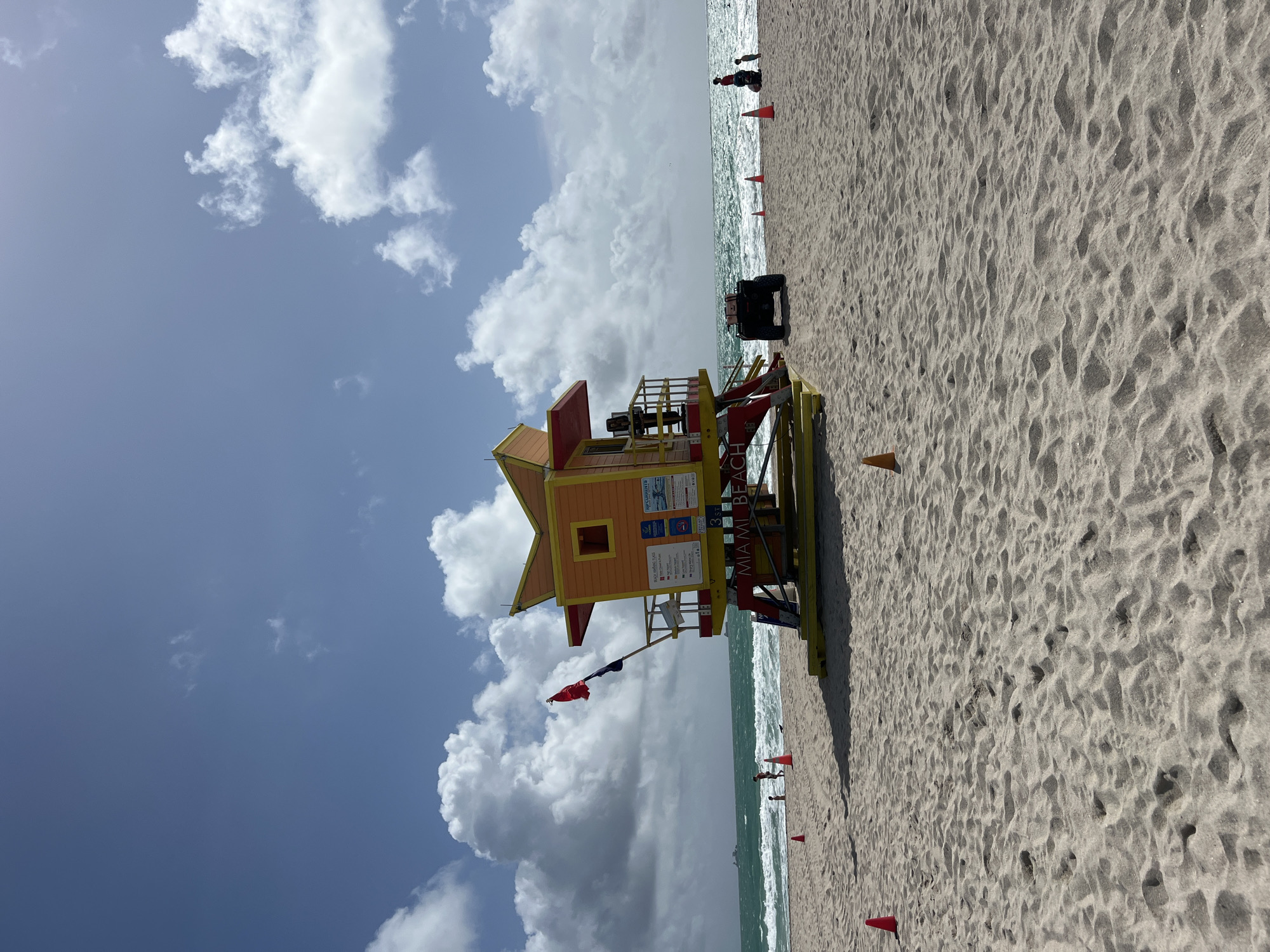 The image shows a lifeguard station on Miami Beach, United States. The station is painted in yellow, orange, and red colors with a star-shaped facade. The beach is sandy, and there are several red cones placed along the shoreline. There is an all-terrain vehicle on the sand near the lifeguard stand. A few individuals are visible in the water and along the shoreline. The sky is blue with scattered white clouds.