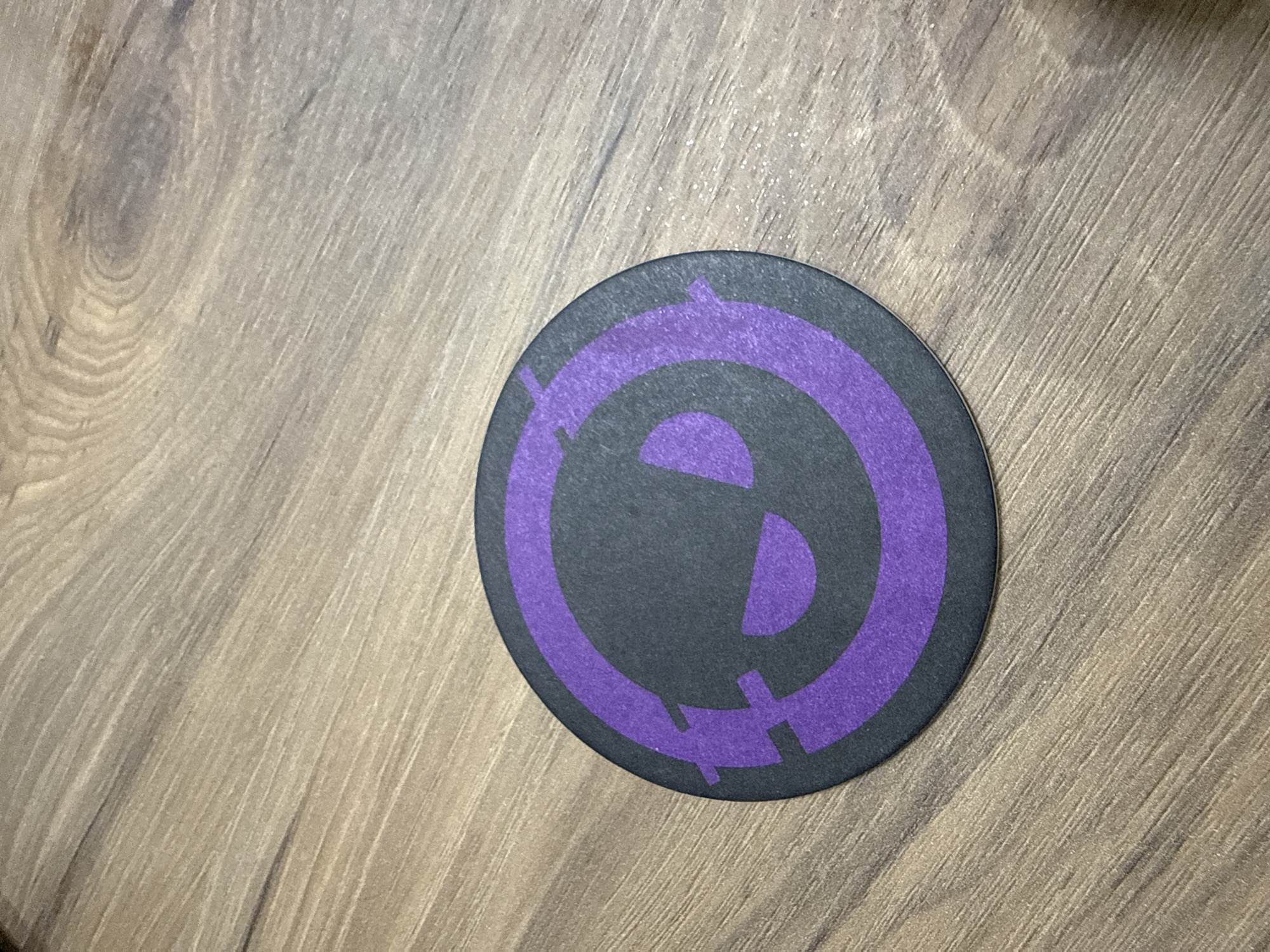 A round, black coaster with a purple design sits on a light brown wood surface. The purple design features two concentric circles, the inner one forming a stylized shape with angular cutouts. There are no people, actions, or interactions visible. The environment is a simple, indoor setting. The location is not discernible from the image.