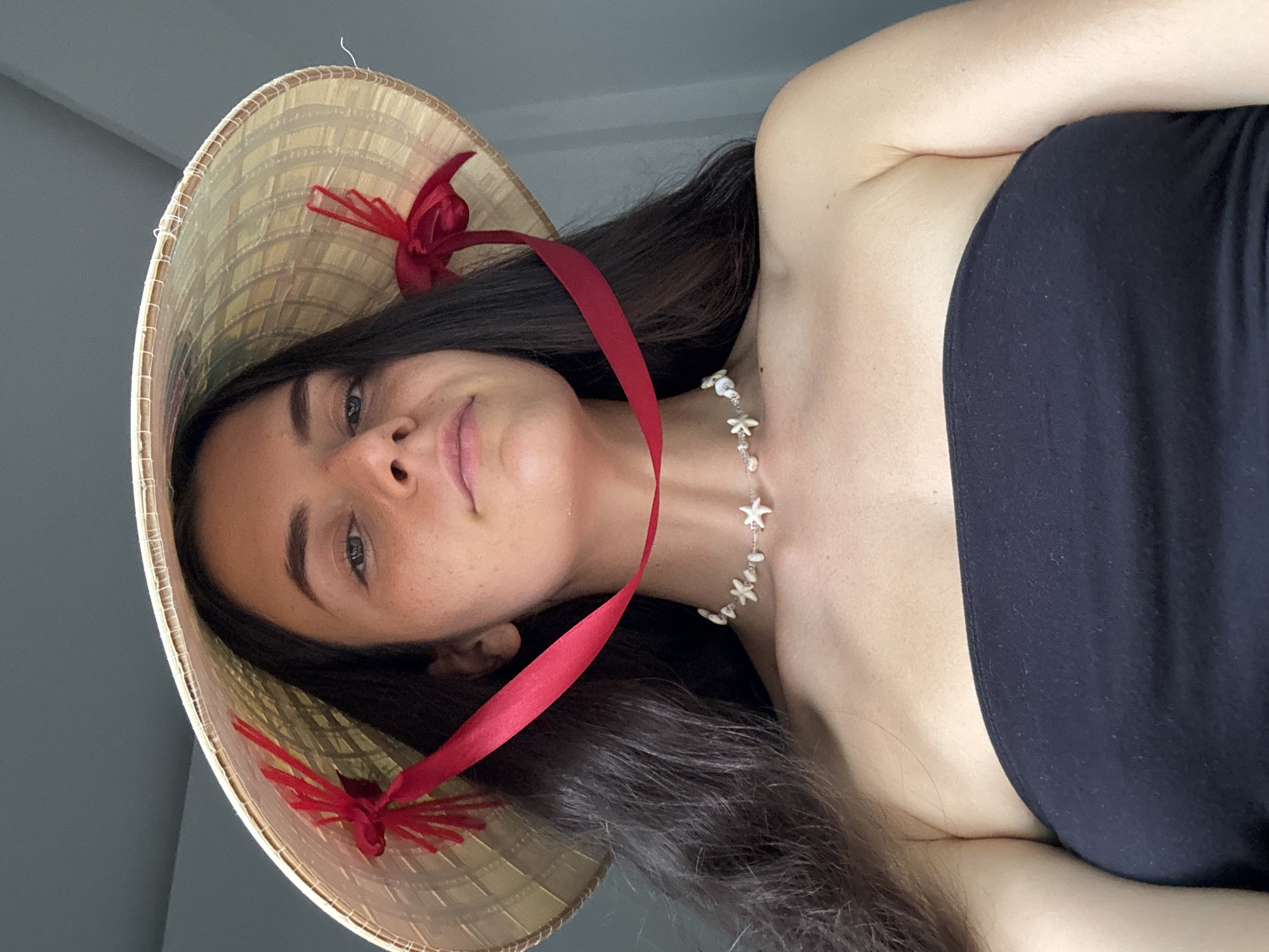 A medium-shot photograph shows a woman wearing a conical Asian hat with a red ribbon and decorations. She is also wearing a black strapless top and a necklace with shell-like pendants. The location is not discernible, and there is no verifiable evidence linking it to Mũi Né, Vietnam. The background appears to be a plain wall. The woman is looking towards the camera.