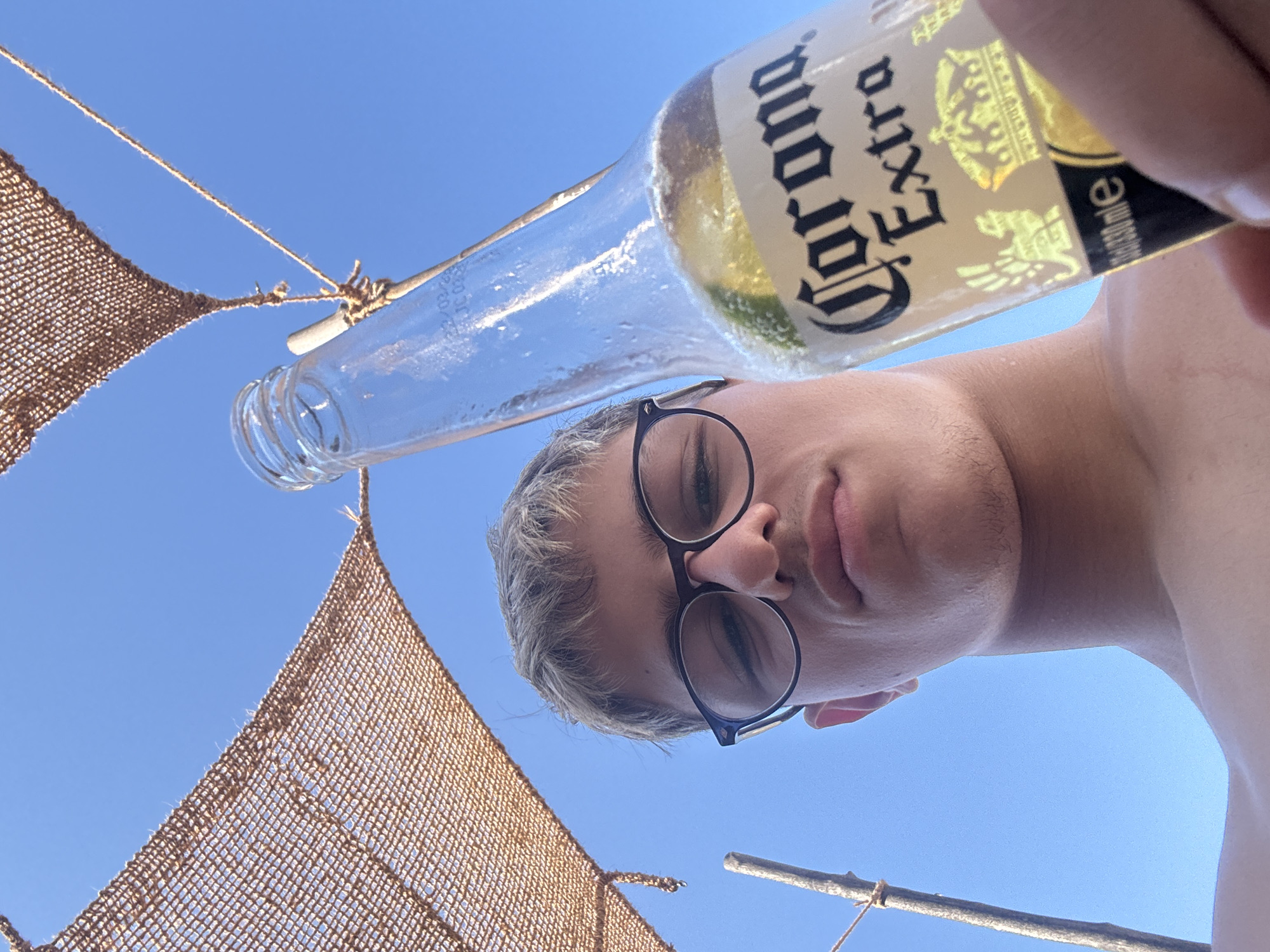 A low-angle, outdoor, close-up shot features a young man holding a bottle of Corona Extra beer against a clear blue sky. He is wearing dark-rimmed glasses and has short, light-colored hair. Part of a woven, brown sunshade is visible in the upper left corner, attached to a wooden pole. The beer bottle, with a lime wedge inside, is held in the foreground, obscuring some of the man's face and body. The scene suggests a relaxed, possibly beach or outdoor recreational setting. The image does not contain any specific identifiable landmarks of Primorsko, Bulgaria.
