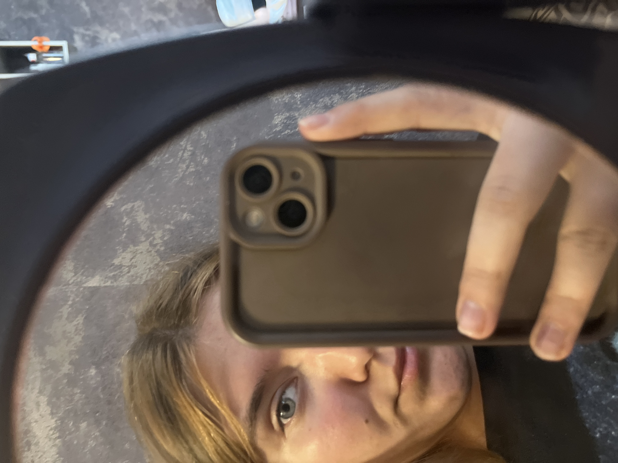A young woman with blonde hair is taking a selfie in a mirror. She is holding a smartphone in her right hand, which is partially obscuring her face. Her left eye and part of her nose and mouth are visible. The background appears to be a textured wall. There are no discernible elements in the image that would allow for the determination of the location within the city of Chaikovsky, Russia.