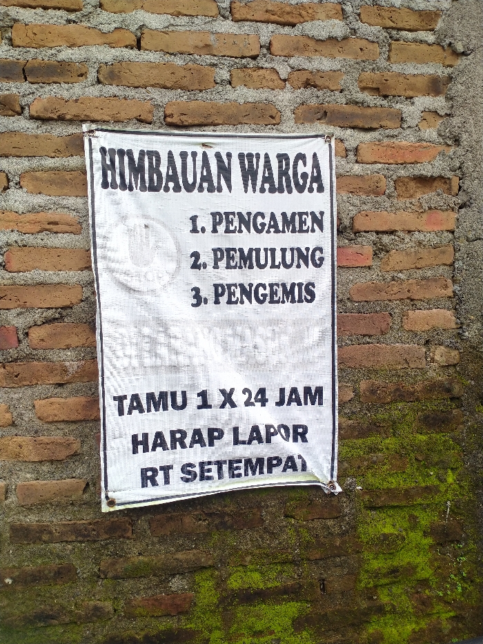 A white sign with black, bold text is prominently displayed against a weathered brick wall. The sign, made of a fabric-like material, is attached to the wall, possibly by nails or staples, and shows some creasing and slight wear. The background is a rustic brick wall composed of reddish-brown bricks with visible grey mortar, indicating an outdoor setting. Patches of vibrant green moss grow on the lower and right sections of the brick wall, suggesting a humid environment or shade.
The text on the sign is in Indonesian and reads:
"HIMBAUAN WARGA" (COMMUNITY ANNOUNCEMENT / WARNING)
"1. PENGAMEN" (1. STREET MUSICIANS/BUSKERS)
"2. PEMULUNG" (2. SCAVENGERS/RECYCLERS)
"3. PENGEMIS" (3. BEGGARS)
Below this list, there's another directive:
"TAMU 1 X 24 JAM" (GUESTS FOR 1 X 24 HOURS)
"HARAP LAPOR" (PLEASE REPORT)
"RT SETEMPAT" (TO THE LOCAL RT - Rukun Tetangga, a neighborhood association leader)
The sign serves as a set of community rules or a warning to residents and visitors in the Kecamatan Kebakkramat, Indonesia, indicating that the presence or activities of street musicians, scavengers, and beggars are being regulated or discouraged. It also stipulates a requirement for guests staying longer than 24 hours to be reported to the local neighborhood head. The overall impression is one of community organization and an effort to maintain order or security within the area. The image appears to be taken during the daytime, with soft, diffused lighting.
