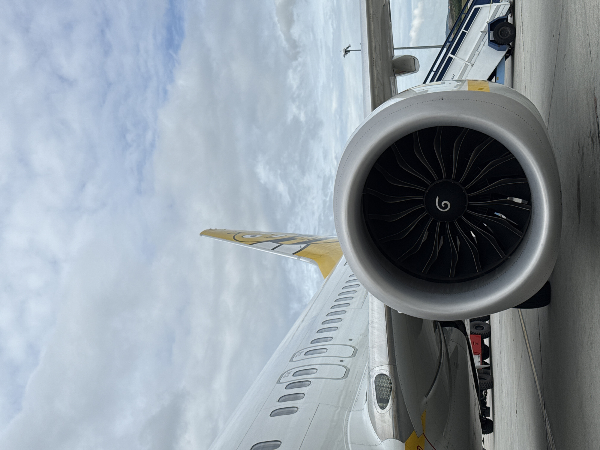 An exterior, eye-level, medium shot captures a close-up view of the left engine of a white and yellow airplane parked on an asphalt tarmac. The massive silver engine cowling dominates the foreground, with its black, swirling fan blades visible within. Behind the engine, the white fuselage of the airplane stretches out of frame, showing a row of oval windows and two larger rectangular doors. The tail of the aircraft, adorned with a yellow and black cartoonish character, is visible on the right. To the right of the airplane, a white and blue mobile staircase is positioned near the fuselage. In the background, a red tug vehicle with large black tires is partially visible. The scene is under a cloudy, overcast sky. The location is identified as the city of Aksakovo, Bulgaria.