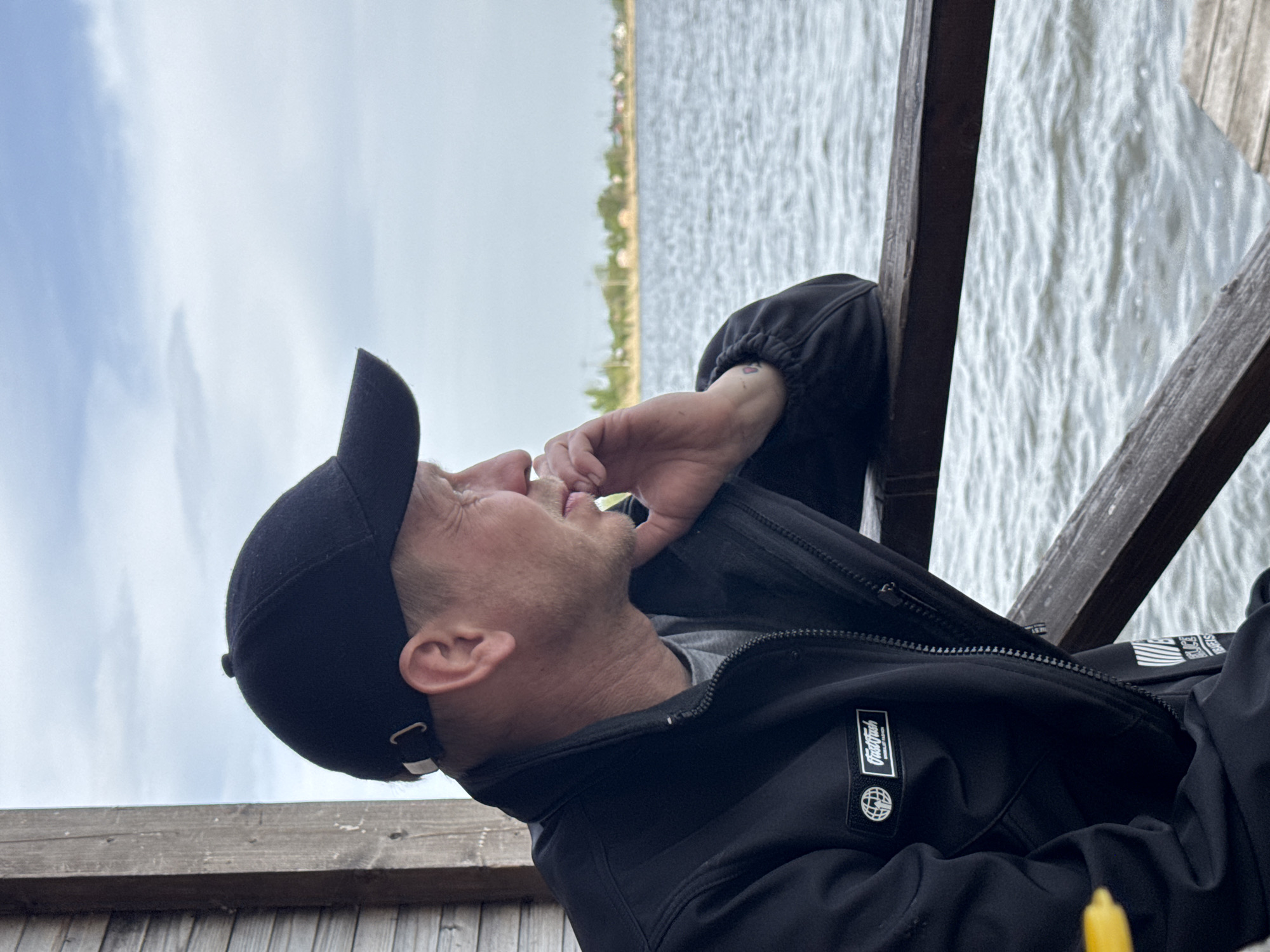 A medium shot depicts a person in Unknown, Unknown, in an outdoor setting. The individual, wearing a black baseball cap and a black jacket, leans against a wooden structure while looking up and to the left. Their right hand is near their face. In the background, a body of water extends to a distant shoreline with sparse vegetation. The sky is overcast.