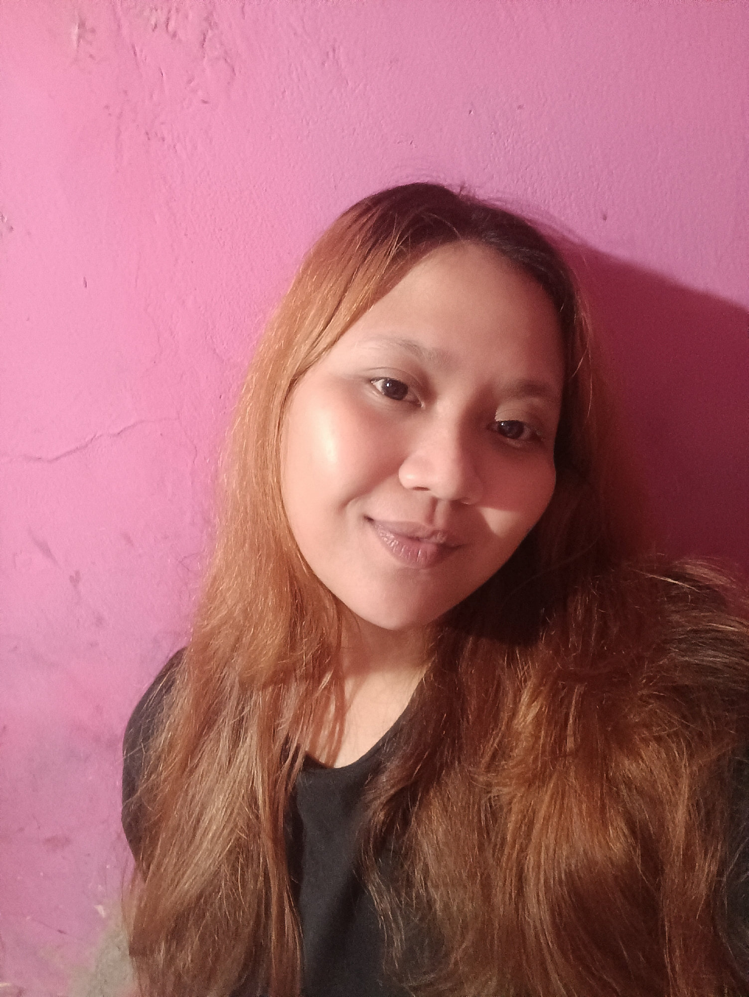 A woman with long, reddish-brown hair is taking a selfie against a bright pink wall. She is wearing a black shirt and is positioned in the center of the frame, with her face angled slightly to the left. The wall behind her appears textured and is lit from the side, casting a shadow on the right side of the image. The lighting suggests an indoor setting. The image is taken from a medium close-up perspective.