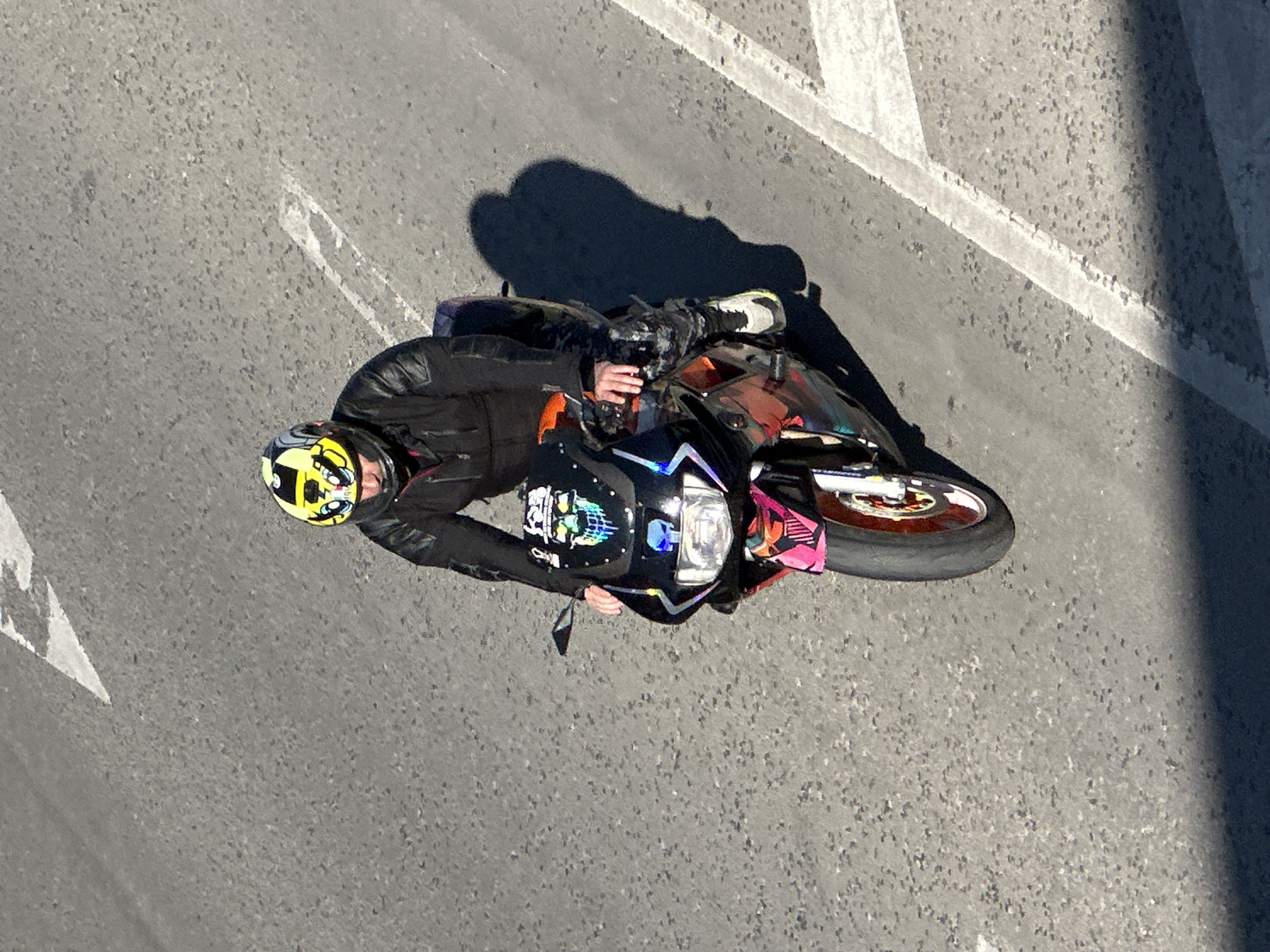 A person is riding a motorcycle on a paved road, likely in Veliko Tarnovo, Bulgaria, given the context. The rider is wearing a black motorcycle jacket and pants, and a yellow helmet with black and white graphics. The motorcycle is black with colorful graphics, including skulls on the front fairing. The rider's shadow is cast on the road to the right. White road markings, including a directional arrow, are visible on the asphalt. The lighting suggests it is daytime, with strong sunlight creating distinct shadows. The environment appears to be an urban or suburban road.