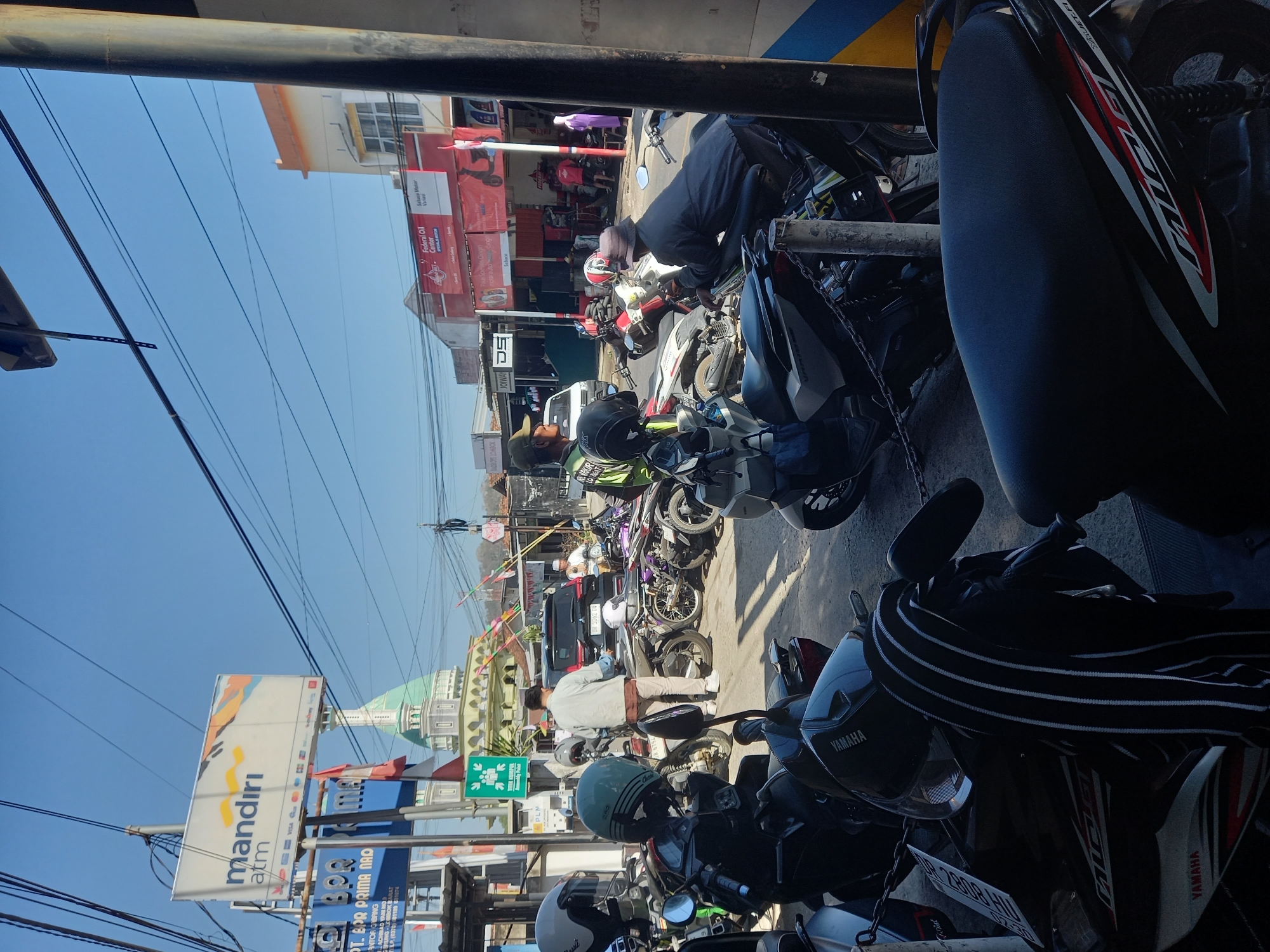 The image depicts a street scene in Gerung, Indonesia, with a focus on a parking area for motorcycles. In the foreground, several parked motorcycles are visible, some with helmets on them. A person in a grey shirt and light-colored pants stands near a parked motorcycle, interacting with another person wearing a hat. In the mid-ground, more motorcycles are parked, and some people are standing around them. A black SUV is parked further back. A prominent Mandiri ATM sign is visible on the left side of the image, along with other shop signs. In the background, a building with a green dome, likely a mosque, stands against a clear blue sky. The street is paved, and power lines are strung overhead. The overall scene suggests a busy, everyday street environment with a concentration of motorcycle activity.