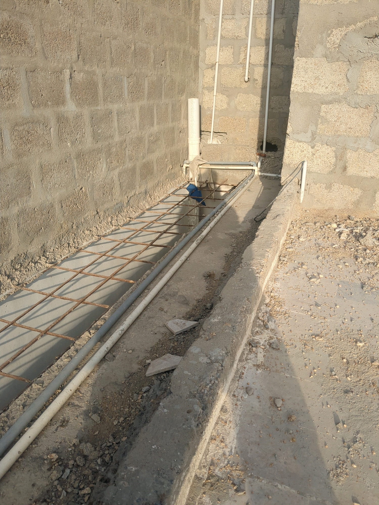 The image depicts a construction site, likely a drainage channel or foundation area, with concrete block walls and a concrete trench. A metal mesh reinforcement grid is laid within the trench, partially covered by a thin layer of concrete. Two plastic pipes, one white and one grey, run parallel to each other within the trench. Several pieces of debris, including small stones and what appears to be a broken tile, are scattered around the trench and on the surrounding ground. A light blue cloth or bag is visible near the metal grid. The environment suggests an arid or dusty location, consistent with areas in Karachi Division, Pakistan, given the construction materials and ground cover. No individuals are present in the frame.