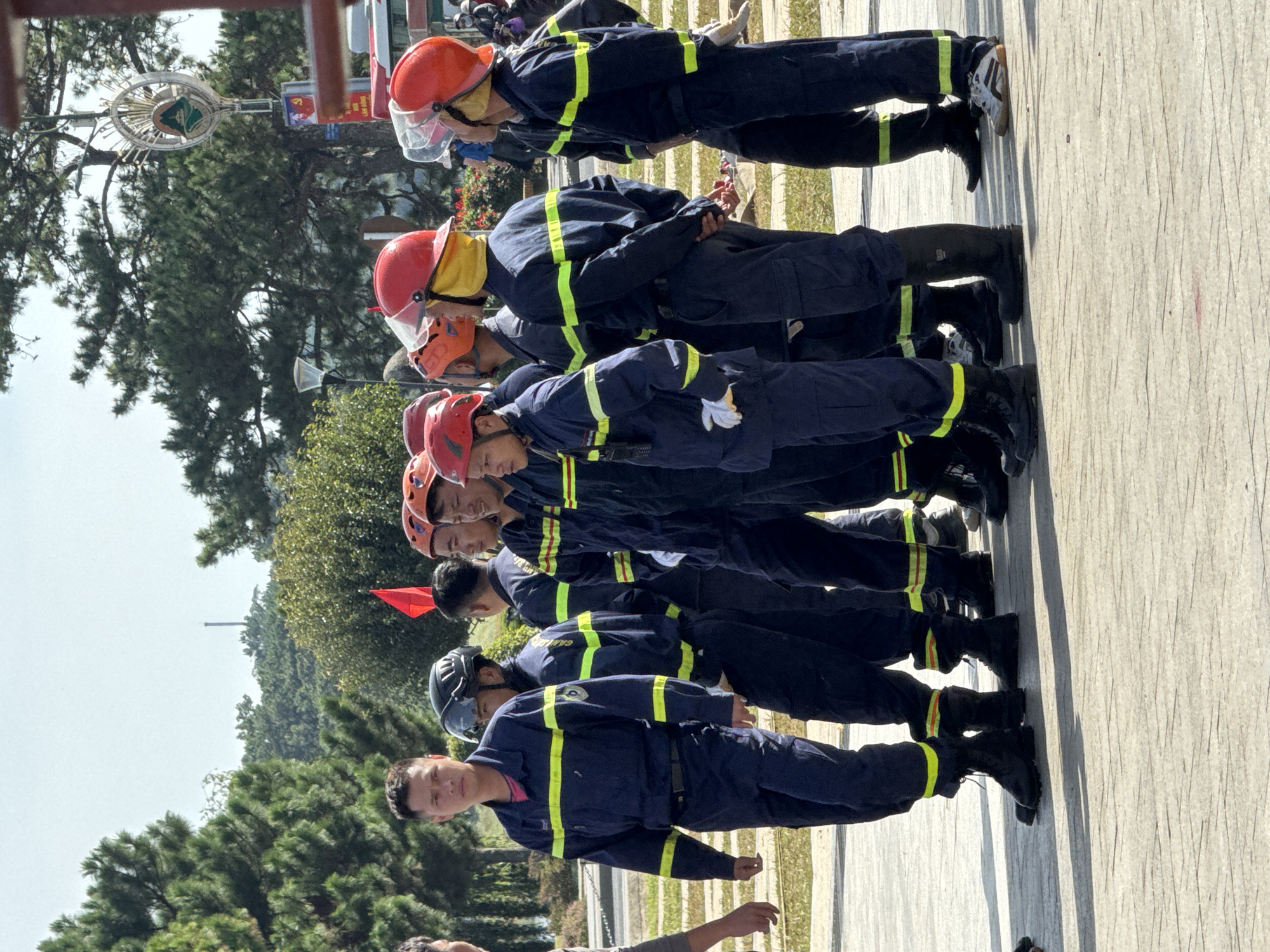 A group of male firefighters stand in formation outdoors. They are dressed in dark blue uniforms with bright yellow reflective stripes on the sleeves and legs, and most are wearing protective helmets in red or orange. The firefighters are standing on a paved surface with a textured pattern. In the background, there are tall pine trees, some foliage, and a clear sky. To the right, a red flag is visible. A partial view of a vehicle and a street sign are also present in the background, suggesting an urban or park setting. The overall impression is one of an organized gathering or training exercise. The presence of the street sign with Vietnamese text and the surrounding vegetation is consistent with a location in Da Lat, Vietnam.