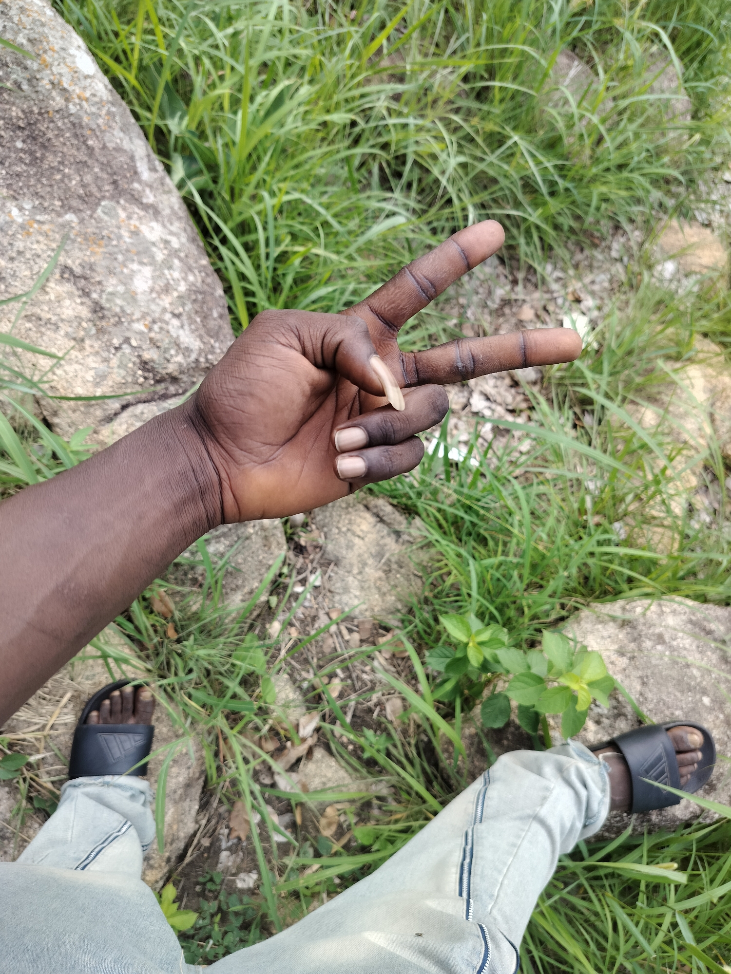 A person's left arm and legs are visible from a top-down perspective, positioned on the ground amidst green grass and rocks. The arm is extended, with the hand held up in a peace sign, fingers bent to form a 'V' shape. The person is wearing light-colored jeans and black slide sandals. The environment appears to be a natural outdoor setting. No specific location in Du, Nigeria can be identified from the image.