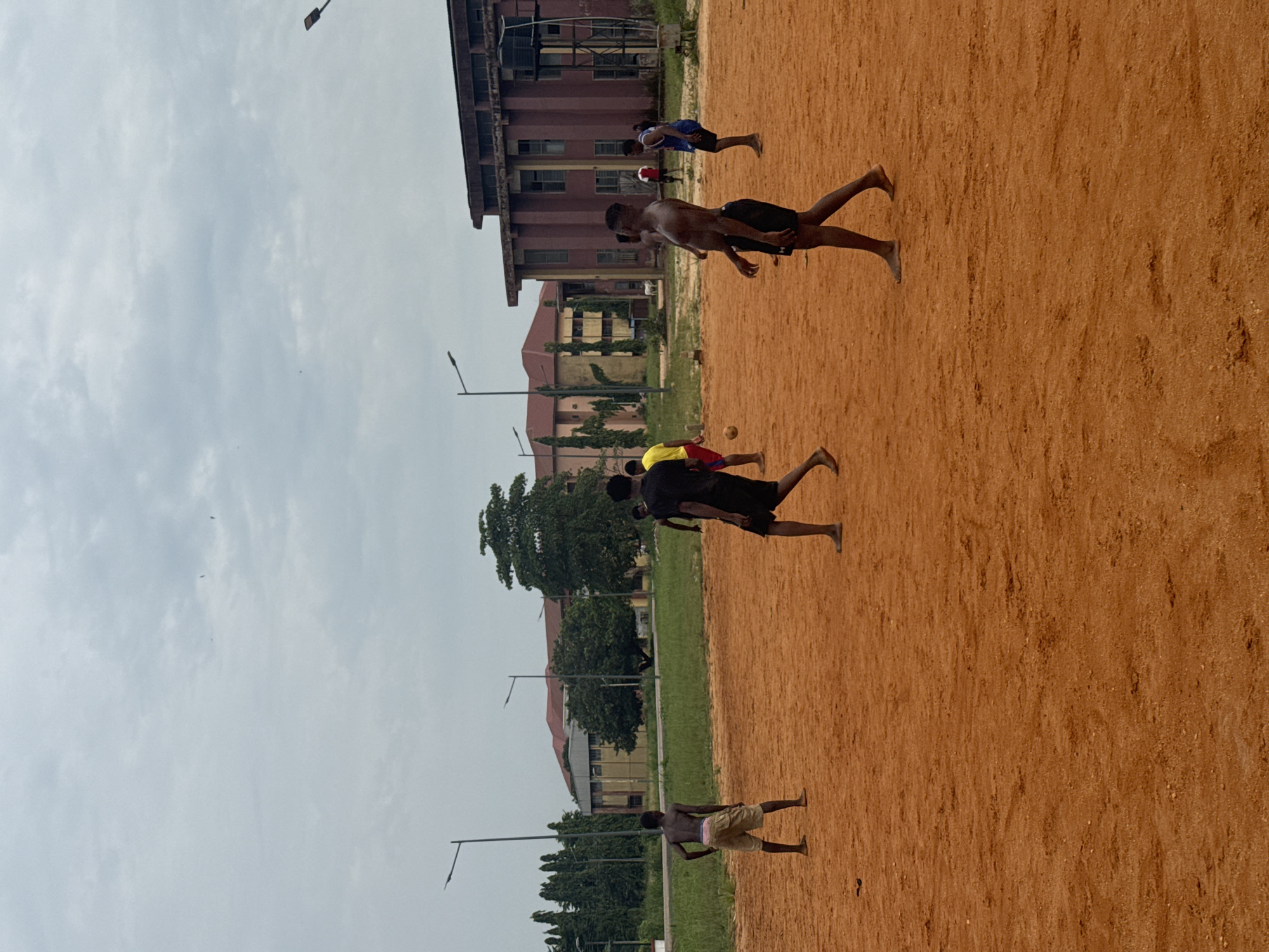 A group of young men are playing soccer on a dirt field in front of several buildings. The sky is overcast. The men are wearing shorts and are barefoot. Some buildings appear to be educational or administrative, with multiple stories and large windows. The field is uneven and reddish-brown, suggesting it is made of laterite soil, common in this region of Nigeria. In the background, there are trees and streetlights. The overall scene depicts a casual sporting activity in an outdoor campus environment.
