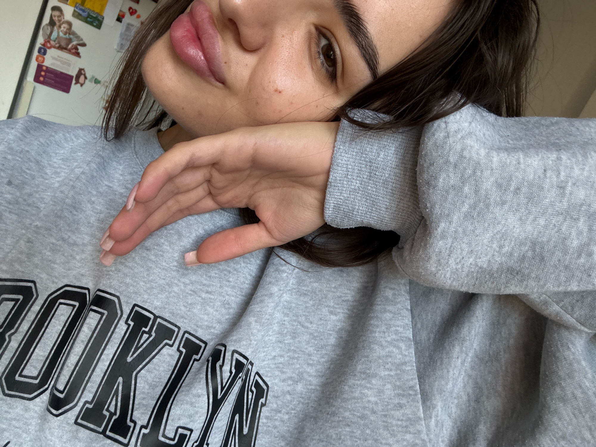 A young woman with dark hair and light skin rests her head on her hand. She is wearing a gray sweatshirt with the word "BROOKLYN" in black letters. Her eyes are looking away from the camera, and her lips are slightly parted. The background is blurry but appears to be an indoor setting. There is no specific information or visual cues that would allow for the determination of the location within the city of Chișinău, Moldova.