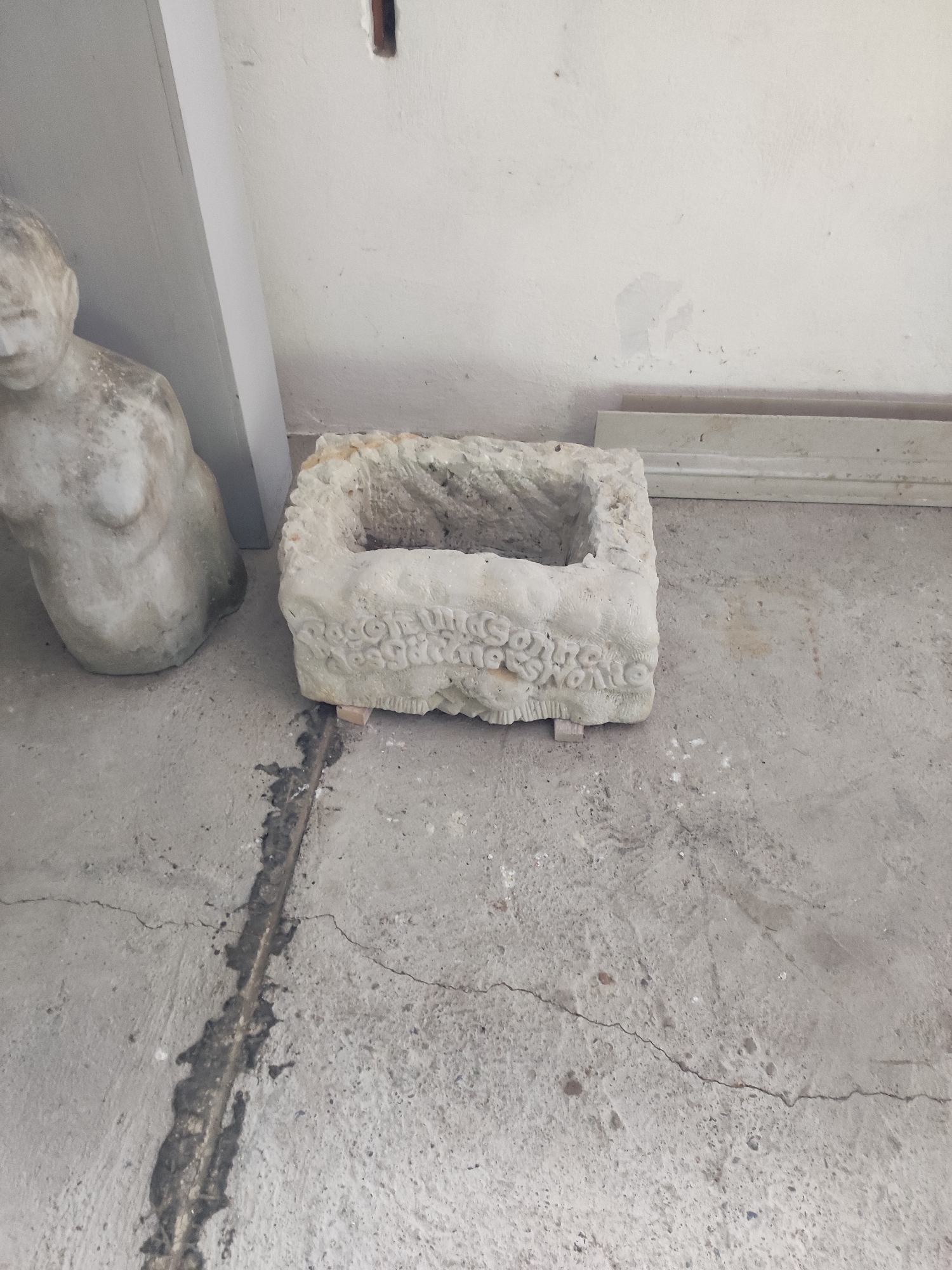 The image shows a rectangular, carved stone vessel resting on a concrete floor. The vessel has an opening at the top and decorative carvings along its sides. The carvings appear to be in relief and depict floral or scroll-like patterns. The stone is light in color and shows signs of age and wear, with some areas appearing rough or pitted. The vessel is slightly elevated by two small, light-colored wooden blocks.
To the left of the vessel, a portion of a light-colored statue is visible. It appears to be the lower torso and hips of a human figure, possibly female, rendered in a similar stone material. The statue is smooth in some areas and has a textured, granular surface in others, suggesting it is also weathered.
The floor is made of concrete and shows cracks and some darker stains, indicating it is likely an outdoor or utilitarian space. A narrow, dark, filled-in channel runs across the floor in front of the vessel. In the background, a white wall is visible, with some discoloration and marks. A vertical white molding or beam is also present on the left side of the image, separating the wall from a darker area.
The scene appears to be indoors, possibly in a storage area, a workshop, or an exhibition space, given the presence of the statue and the utilitarian nature of the floor and wall. There is no clear indication of the time of day or weather. The objects are static, and no activity is taking place. No visible text is present in the image.