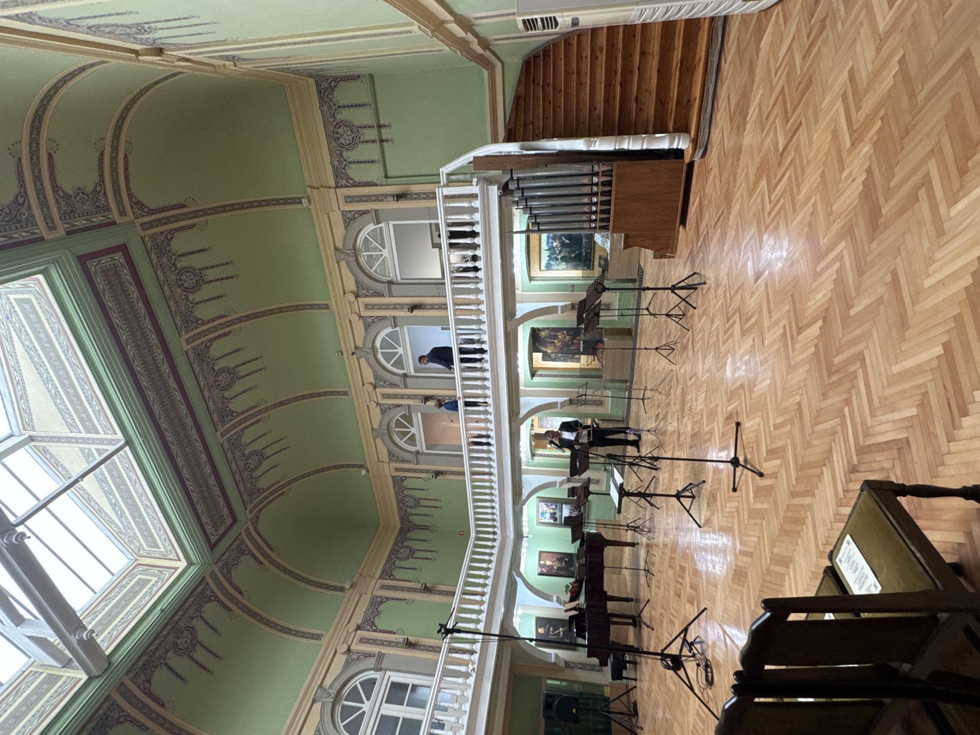 The image shows an interior view of the National Revival Hall within the Regional Ethnographic Museum in Plovdiv, Bulgaria. The room has green walls adorned with decorative paintings. The floor is herringbone patterned parquet. A balcony with a white railing runs along the upper level. On the balcony, two people can be seen. On the main floor, there are two pianos set up, along with multiple music stands. A musician appears to be standing near one of the pianos. Some objects are chairs, some stands, and paintings on the walls. Natural light enters through a skylight in the ceiling. A pipe organ is also visible next to a wooden staircase.