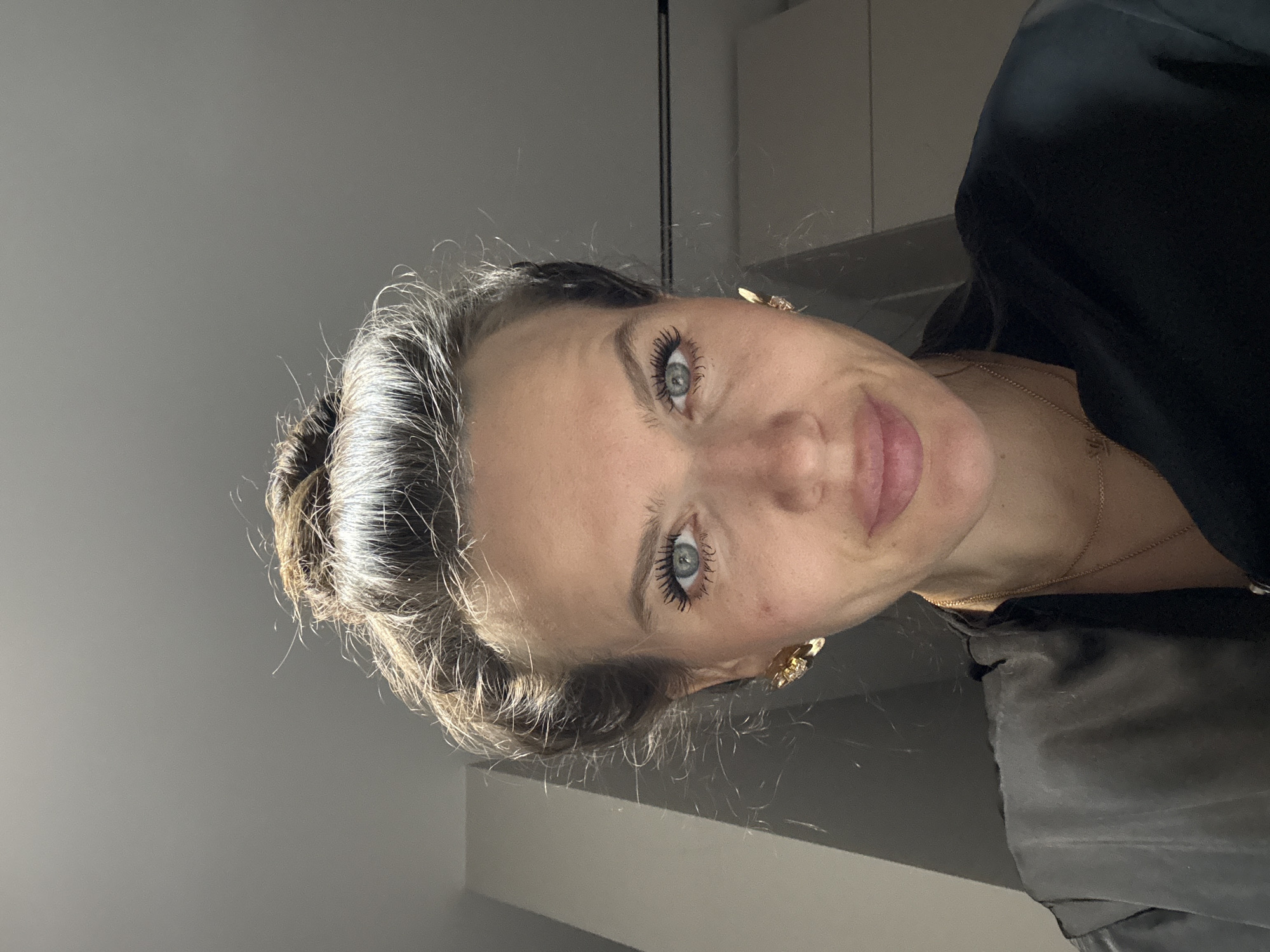 The image shows a woman with light skin, blue eyes, and dark hair pulled back in a bun. She is wearing gold earrings and a necklace. She is dressed in a dark-colored top. The background is a plain, light gray wall. The location is unknown.