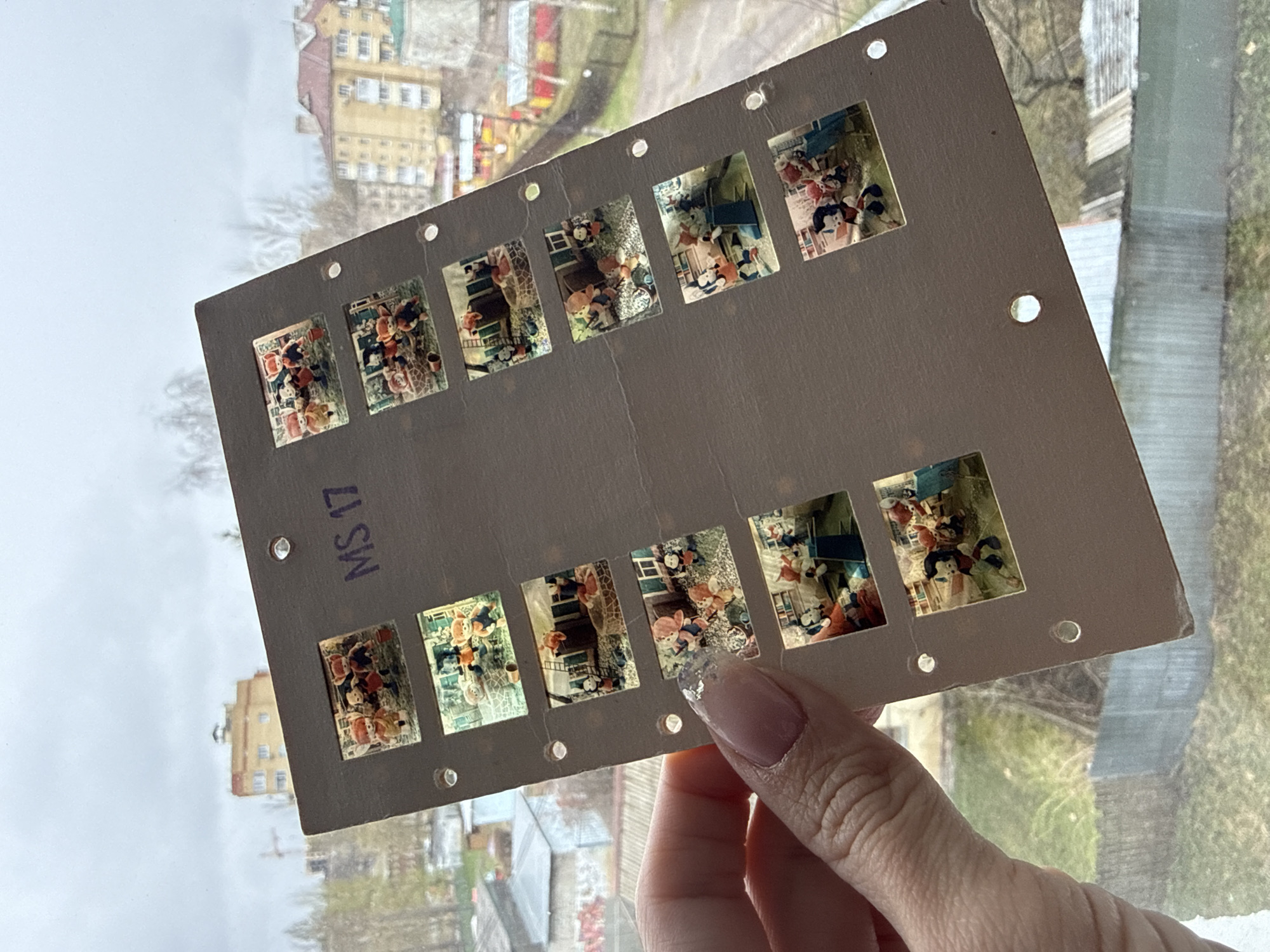A hand holds a strip of mounted photographic slides, with ten visible images of dolls arranged in outdoor scenes. The strip is cardboard, tan or grey, with symmetrically placed circular holes along the edges. The slides show scenes depicting the dolls in various outdoor settings, some with dollhouse-like structures. Text reading "MS 17" is visible on the cardboard. The background is a view of a city, likely Rusanovo, Russia. Visible are buildings, trees without leaves, and rooftops, suggesting an overcast day. A windowpane and its reflection are also visible behind the strip.