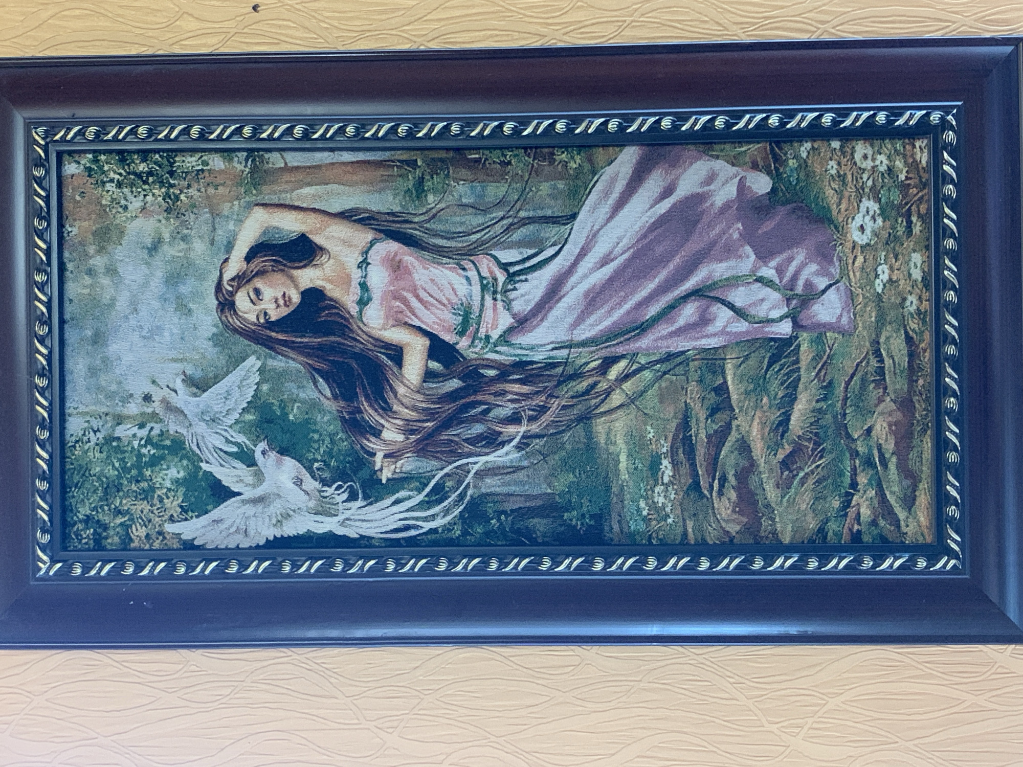 A framed tapestry depicts a woman with long, flowing dark hair and a pink dress standing in a forest. Two white birds with outstretched wings are in flight above and to the left of the woman. The woman is looking towards the viewer with her right hand touching her head. The tapestry is enclosed in a dark brown frame with ornate gold detailing around the inner edge. The background of the tapestry features trees, foliage, and rocks. The tapestry is hanging on a textured, pale yellow wall.