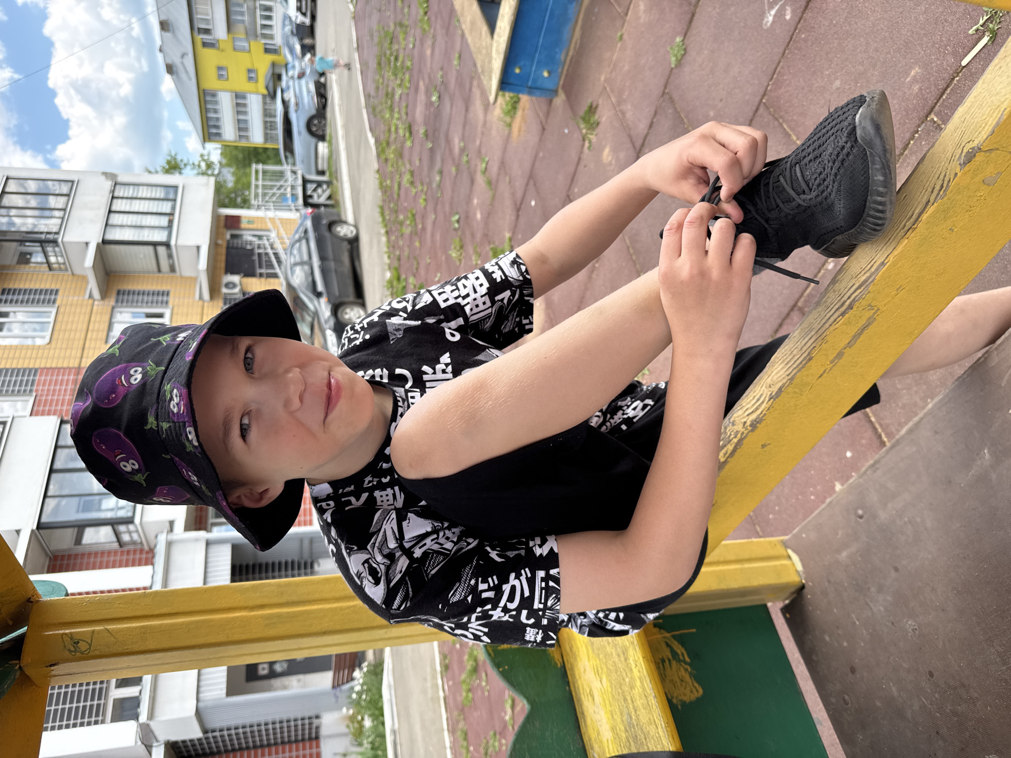 A young Caucasian male, appearing to be around 10-12 years old, is shown outdoors on a playground. He is wearing a black bucket hat with a purple eggplant pattern and a black t-shirt with a white graphic print. He is sitting on a yellow wooden structure, a part of the playground equipment, and is in the process of tying his black shoelaces on his black sneakers. His left leg is bent and resting on the yellow beam. The background reveals a residential area with apartment buildings and parked cars. The ground beneath him is a reddish-brown surface, typical of a playground, with patches of green grass. The sky is partly cloudy. This scene is situated in the city of Zav'yalovo, Russia.