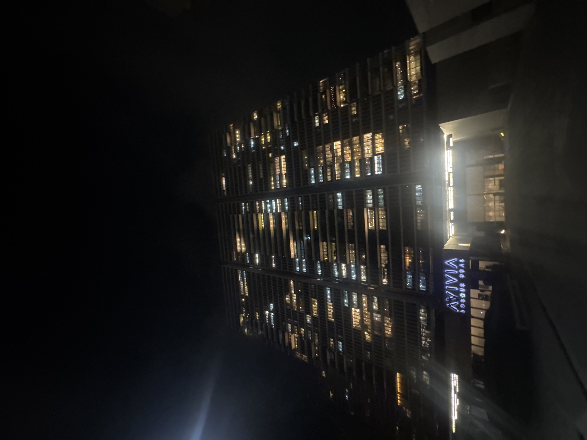 The image depicts the exterior of the hotel "Vivia Hotel" in Huixquilucan de Degollado, Mexico, at night. The building is tall, modern, and brightly lit with numerous windows showing illuminated interiors. The night sky is dark.