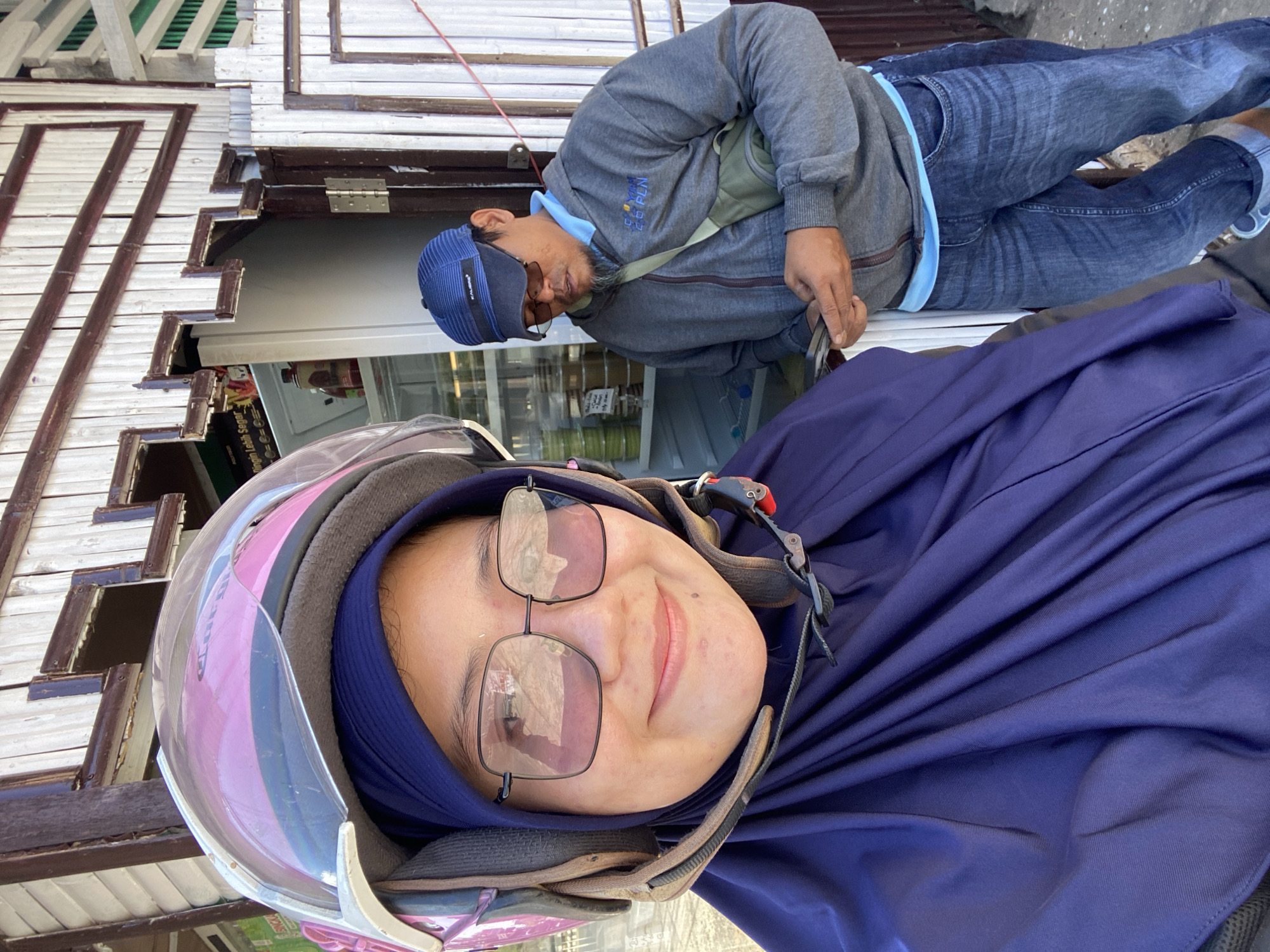 In the city of Sungai Paring, Indonesia, a woman wearing a purple hijab and a pink helmet is taking a selfie. She is also wearing large square-framed glasses. Behind her, a man wearing a blue baseball cap, a gray hoodie with a light blue undershirt, and blue jeans is looking at his phone. He has a small green bag slung across his chest. They appear to be standing in front of a small storefront with a white wooden exterior and a door that is slightly ajar, revealing a refrigerator stocked with items inside. The overall scene suggests an outdoor marketplace or a small retail establishment.