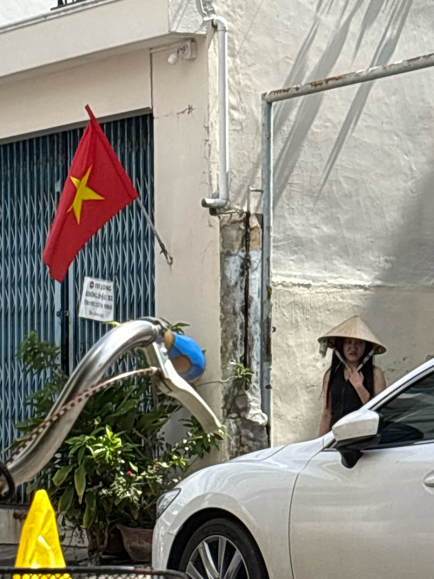 In Nha Trang, Vietnam, a red Vietnamese flag with a yellow star is displayed on a building's exterior, partially obscuring a blue object mounted on a metal pole. Below the flag, a white sign with Vietnamese text is posted. A person wearing a conical hat and a black top is visible on the right side of the image, standing near a white car. The scene is partially lit by sunlight, casting shadows from overhead objects onto the textured wall. In the foreground, a bicycle's handlebars and part of its frame are visible, along with a potted plant and a yellow traffic cone.