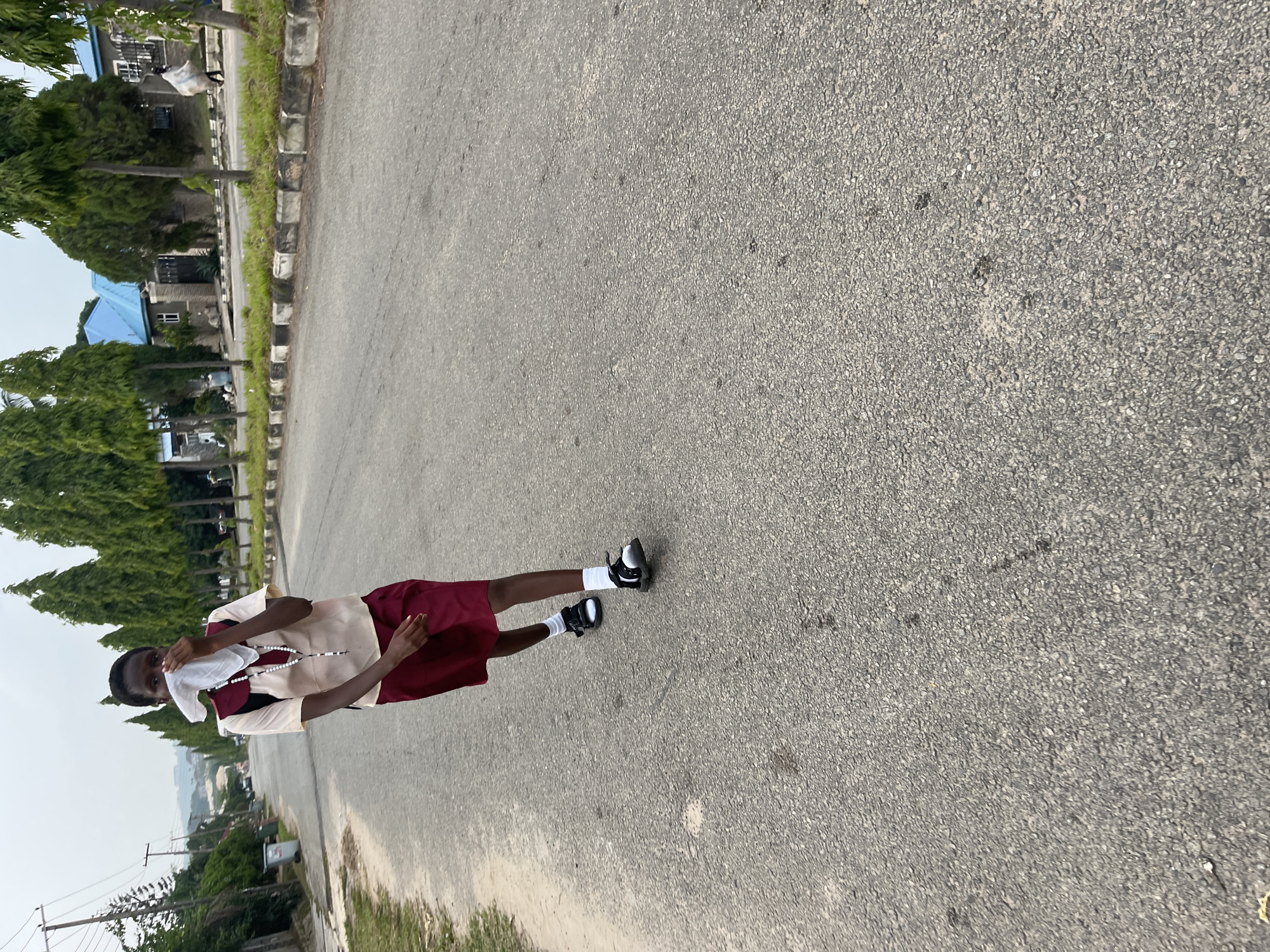 The image, rotated 90 degrees clockwise, captures a young person standing on a paved asphalt road in what appears to be a residential or institutional area in Abuja, Nigeria.
The main subject is a young individual, likely male, positioned slightly to the left of the frame's center. He is dressed in a light beige and maroon top, paired with maroon shorts, white ankle socks, and black sandals. He is facing the camera, holding a white cloth to the lower part of his face, partially obscuring his mouth and nose. A beaded necklace is visible around his neck, hanging over his top.
The setting consists of a wide, grey asphalt road with a textured surface, indicating regular use. To the left of the road, a strip of green grass borders a black and white checkered curb. Beyond the curb, a row of tall, green conifer-like trees lines the street. Through gaps in the trees, several buildings with distinctive blue-colored roofs are visible, suggesting local architectural styles. In the distant background, a hazy landscape with more trees and possibly hills can be discerned under an overcast sky. Utility poles and overhead lines are also visible in the left background.
The activity appears to be a casual pose for a photograph, with the subject looking directly at the camera. The time of day seems to be daytime, likely late morning or afternoon, given the ambient light and lack of harsh shadows. There is no visible text in the image.