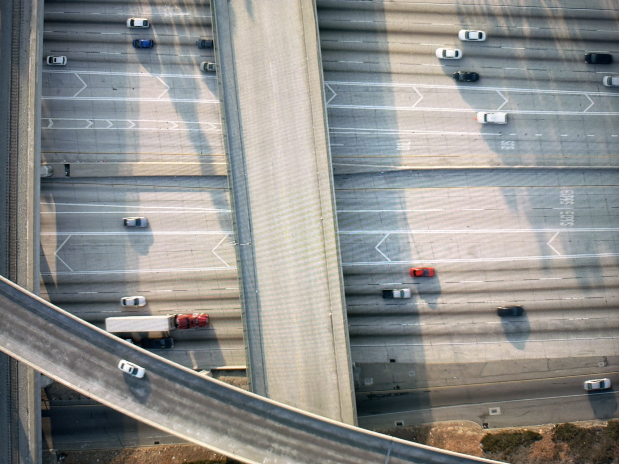 The image is an aerial view of a multi-level highway interchange. Several vehicles are visible on the roadways, including cars, a truck with a red cab and a white trailer, and a bus. The environment appears to be an urban or suburban area with elevated roadways and concrete structures. There are no visible signs or markings indicating the specific location within Kyiv, Ukraine. The lighting suggests it is daytime, with shadows cast by the vehicles and structures. The overall scene depicts a complex transportation network.