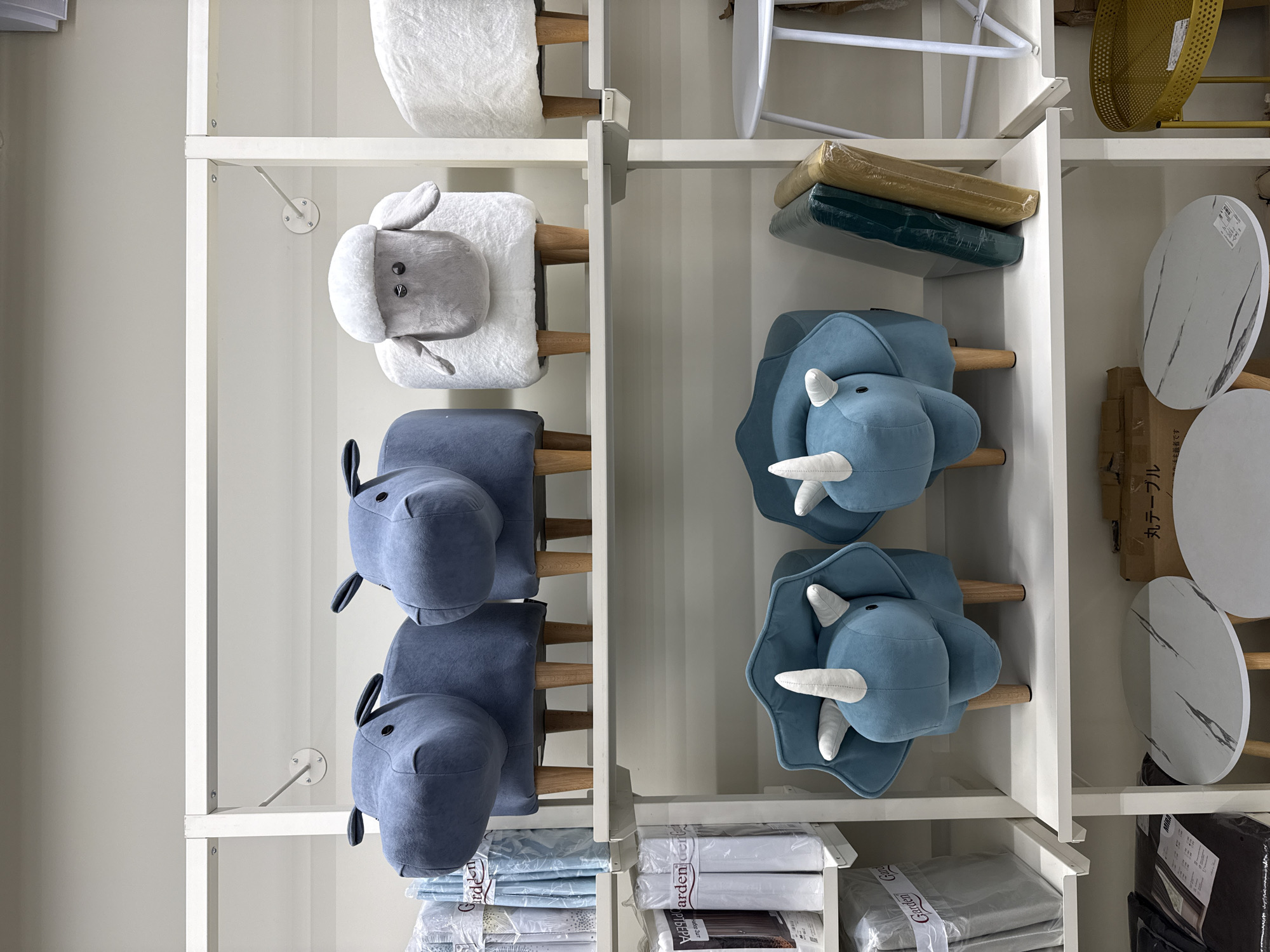 The image depicts a section of shelving within a store, likely located in Izhevsk, Russia, given the presence of text written in a script commonly used in Russia and neighboring regions. The shelving units, painted white, display an assortment of children's stools and household items. The stools are designed to resemble animals; there are stools shaped like sheep, hippopotamuses, and triceratops, all upholstered in plush fabric. Other items on the shelves include packaged textiles, possibly bedding or towels, as well as round marble-patterned tabletops.