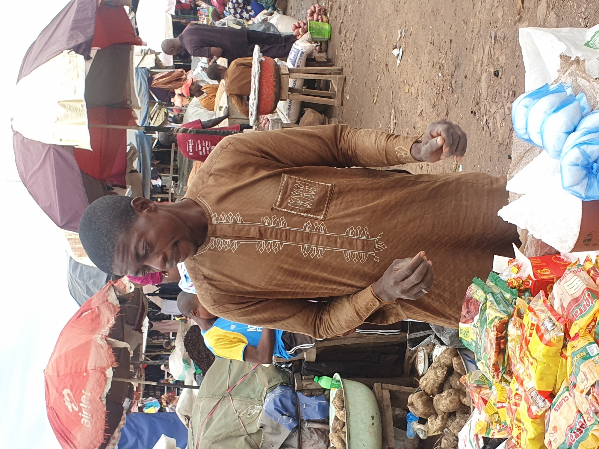 This is a photograph taken at an outdoor market in Lokoja, Nigeria. The image is oriented vertically. In the foreground, a man wearing a brown, embroidered tunic is looking down, his hands clasped in front of him. Behind him, a market scene unfolds with stalls, various goods, and other people.
To the left of the man, a large, red and white umbrella with the "Airtel" logo is visible. Further back, a stall displays bags of colorful snacks or food items, some with text such as "Jerra", "KEN", and "100 CUBES". There are also piles of root vegetables, possibly yams or cassava, in a green basin. Several other people are present, some browsing the stalls and others working. One person in the background wears a dark robe, while another is in a yellow and blue shirt. The ground appears to be dusty earth, typical of an outdoor market. The lighting suggests it is daytime, likely a busy market day. The atmosphere appears to be one of commerce and daily life.