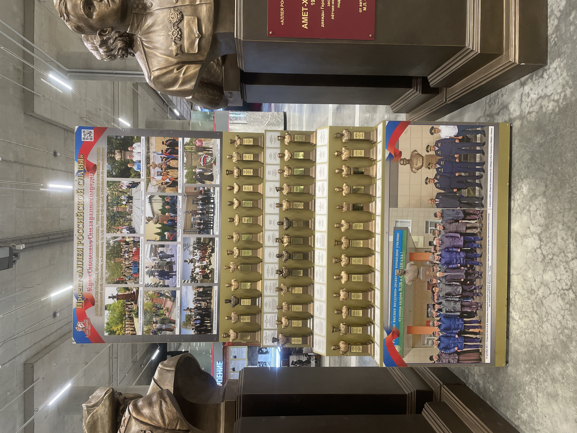 The image shows a museum exhibit, likely in Kronstadt, Russia, dedicated to military heroes. The exhibit features a large display board with photographs of various events and individuals, as well as rows of bronze busts of historical figures. The busts are arranged on tiered stands, with each bust accompanied by a descriptive plaque.
The main display board has the title "Project 'Alley of Russian Glory'" in large lettering. Below this, it states "Heroes of the Soviet Union" and lists names and biographical information. The photographs on the board depict gatherings, ceremonies, and portraits of military personnel and civilians. Some of the photos show busts being unveiled or dedicated.
To the right of the main display, there is another vertical display with a red background, featuring text that reads "«АЛЛЕЯ РОССИЙСКОЙ СЛАВЫ»" and "АМЕТ-ХАН, СУЛТАН", followed by details about his achievements as a twice Hero of the Soviet Union and an honored pilot.
In the foreground and on the sides, two large bronze busts of military figures in uniform are visible, suggesting they are part of the exhibit or flanking the display. The busts are detailed and wear military caps and insignia.
At the bottom of the main display board, a photograph shows a group of people, some in uniform and others in civilian attire, standing in front of a building. A text box below this photo indicates the date and location of the event: "27 декабря 2017 г. Открытие бюста В.В. Маргелова в Рязанском высшем воздушно-десантном командном училище имени генерала армии В.Ф. Маргелова, г. Рязань." (December 27, 2017. Unveiling of the bust of V.V. Margelov at the Ryazan Higher Airborne Command School named after Army General V.F. Margelov, Ryazan).
Another photograph at the bottom of the display shows a group of men, mostly in uniform, standing outdoors. Below this photo, the text reads: "3 августа 2019 г. Открытие памятников А.В. Лебедя и В.А. Грачёву в РВВДКУ, г. Рязань." (August 3, 2019. Unveiling of monuments to A.V. Lebed and V.A. Grachev in RVVDKU, Ryazan).
The environment appears to be an indoor public space, possibly a museum or a gallery, with concrete floors and ceiling, and fluorescent lighting. The overall impression is one of solemn commemoration and tribute to military service and heroism.