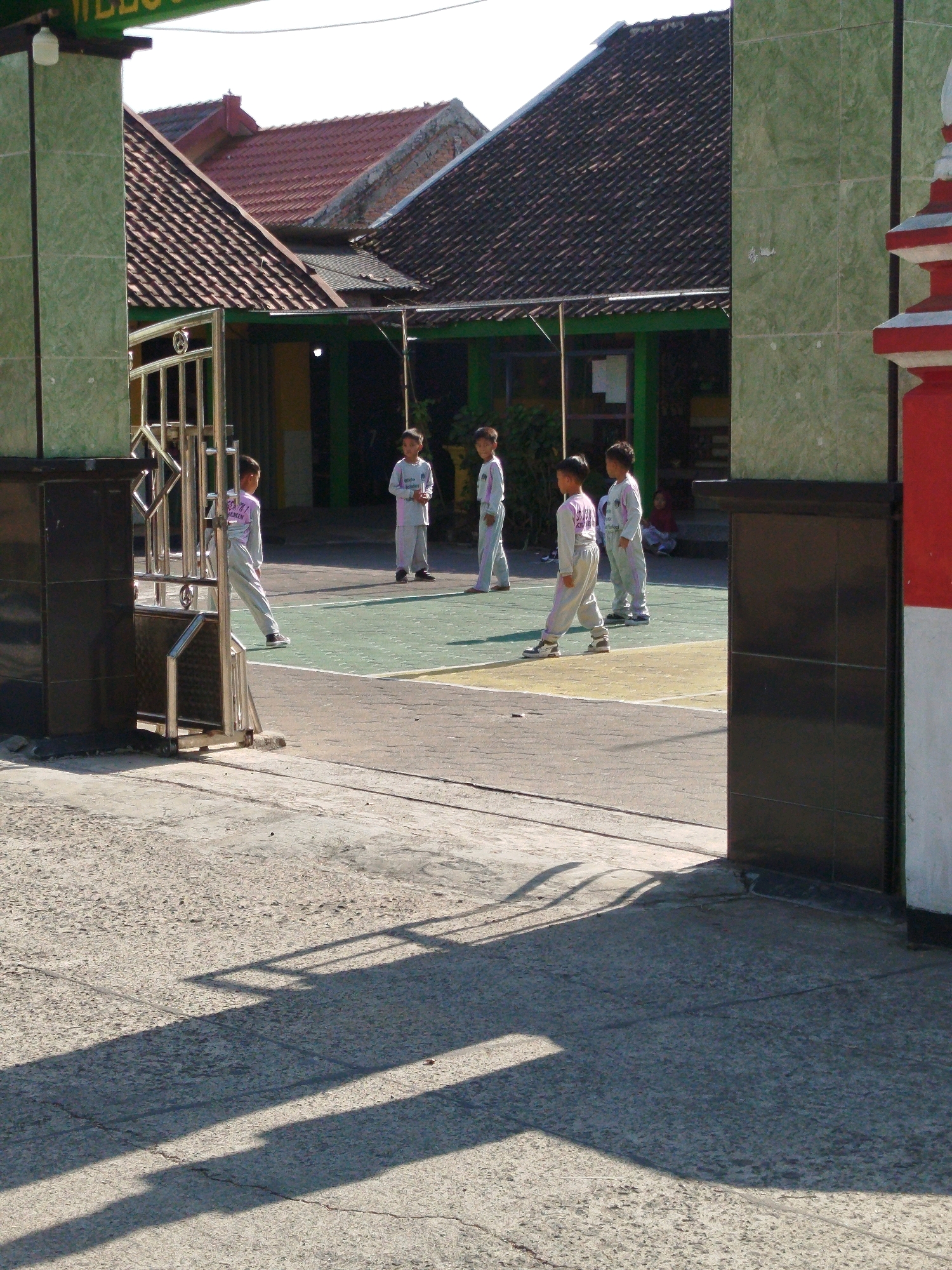 The image shows a group of young students in a schoolyard in Semin, Indonesia. The students are wearing matching white uniforms with purple accents and appear to be standing on a paved area with markings that suggest a sports court. There are several school buildings with red-tiled roofs visible in the background. A metal gate is partially open, allowing access to the school grounds. The lighting suggests it is daytime, with shadows cast on the ground from the gate and surrounding structures.