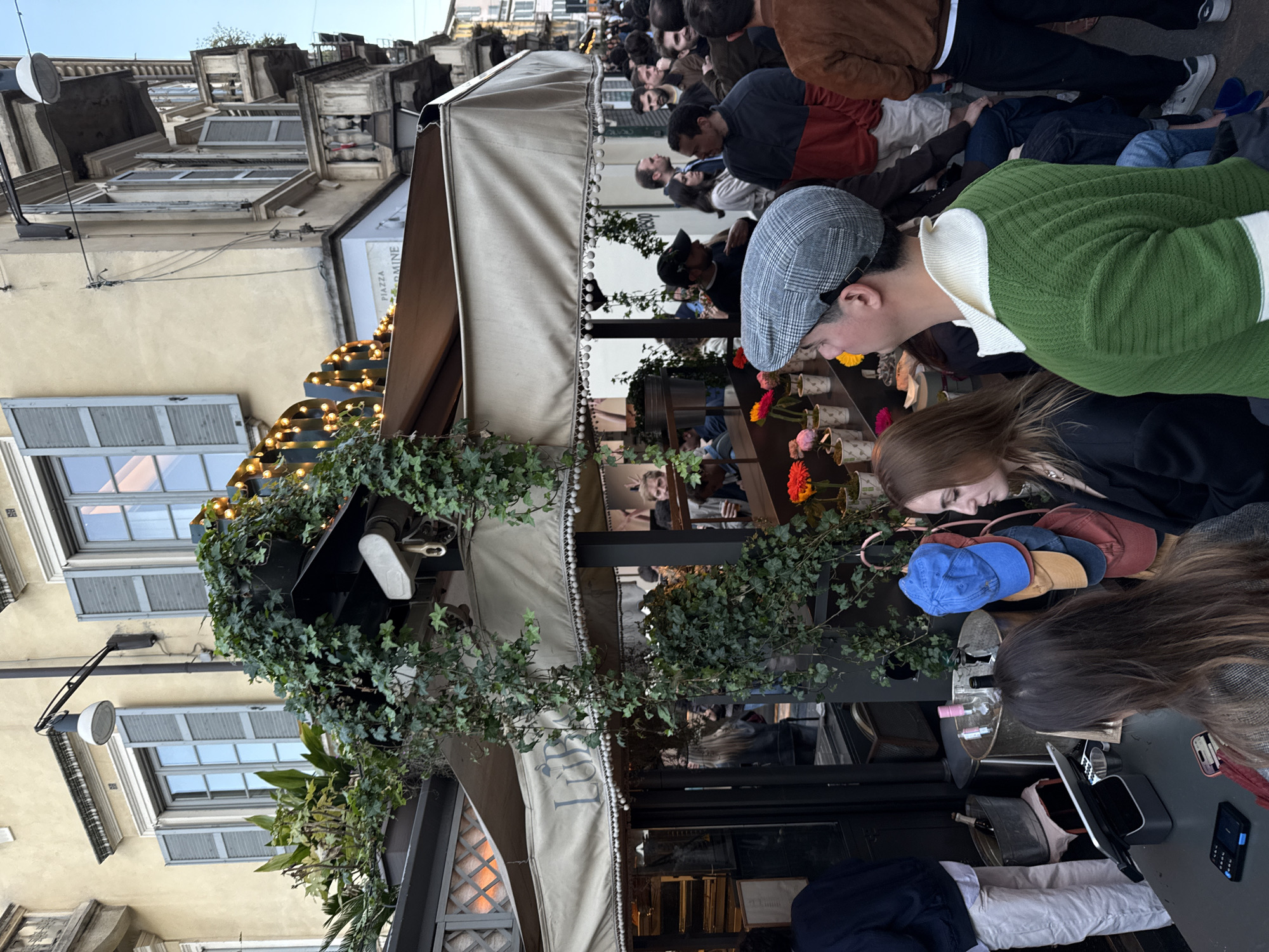 The image shows an outdoor scene in Milan, Italy. A crowd of people is gathered outside a building adorned with awnings, greenery, and string lights. Some individuals are looking at items displayed on a shelf or counter. There are several people seated, including a person wearing a green sweater and a cap. A table is visible, holding items like hats, a bottle, and a telephone. The building has windows with shutters. The scene is vibrant and bustling, suggesting a market or event taking place in the city.