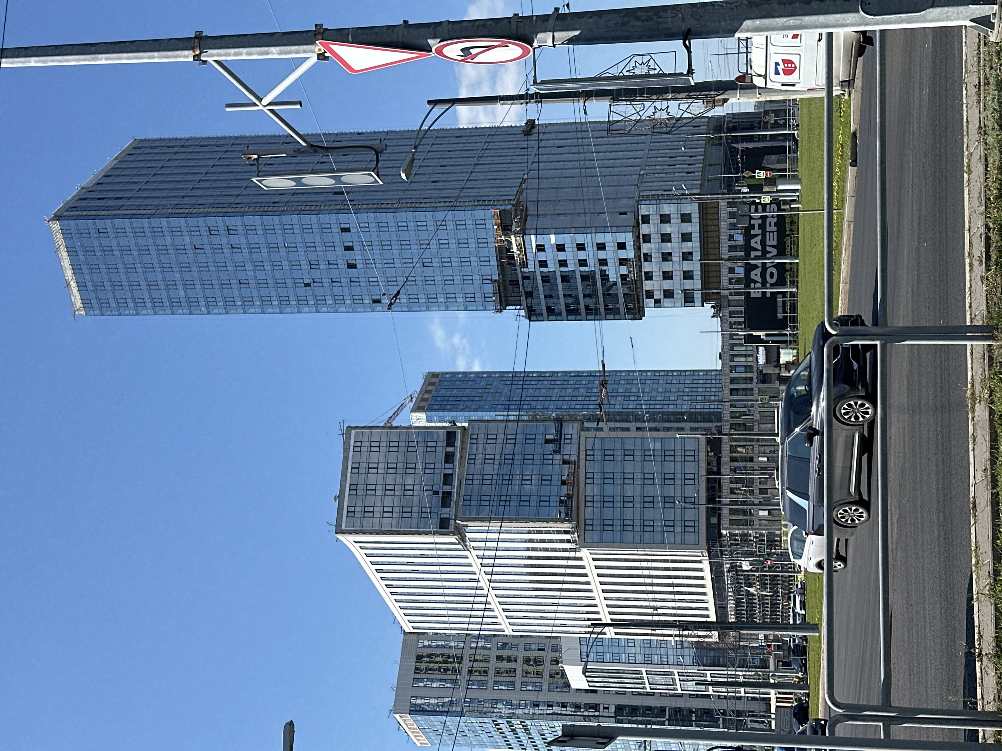 In a bright, clear sky, multiple contemporary skyscrapers dominate the skyline of an urban setting. The buildings are characterized by their sleek, glass facades, reflecting the sunlight. One particularly tall, slender tower stands on the right, its numerous windows creating a grid pattern. To the left, a cluster of interconnected buildings features a more complex geometric design, with stepped sections and varied window arrangements. Construction cranes are visible near the tops of some structures, indicating ongoing development. In the foreground, a multi-lane road is present, with several vehicles including a dark-colored SUV and a white van. The road is bordered by a grassy median with some yellow wildflowers. Streetlights and traffic signals are positioned along the roadside. A large banner displaying text is visible on the lower section of one of the buildings.