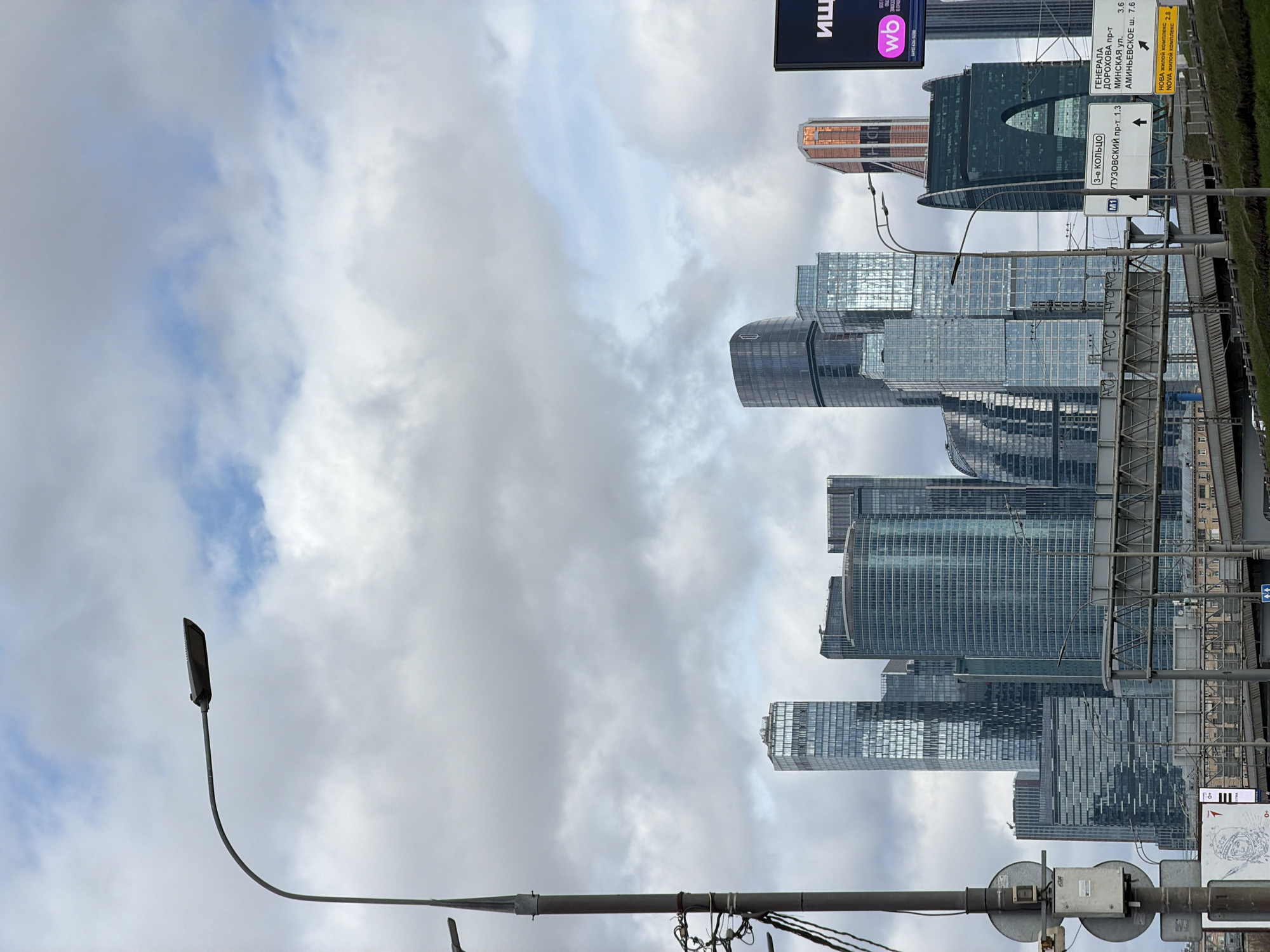 The image shows a cityscape of Moscow, Russia, featuring a group of modern skyscrapers against a cloudy sky. In the foreground, there is a street lamp and various road signs with directions, including indications for the "3-е КОЛЬЦО" (Third Ring Road), "Тузовский пр-т" (Tuzovsky Avenue), and directions to other streets and residential complexes like "Генерала Дорохова пр-т" (General Dorokhov Avenue) and "Аминьевское ш." (Aminevskoe Highway). A sign with "wo" and "Ищ" in Cyrillic is also visible. The presence of skyscrapers, combined with road signs in Russian, indicates that the image was taken in a modern business district of Moscow. The cloudy sky suggests overcast weather conditions. There are no people directly visible in the image.