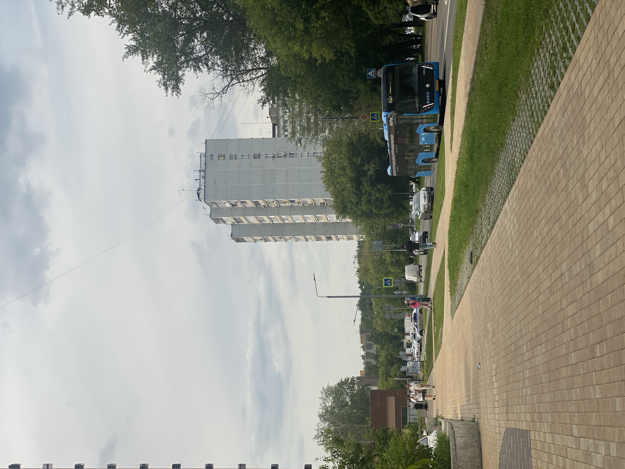 The image depicts a street scene in Moscow, Russia, on a cloudy day. In the foreground, a pedestrian walkway paved with light brown rectangular bricks leads towards the viewer. Adjacent to the walkway is a strip of green grass and a grey gravel surface with hexagonal indentations.
The street itself is paved with asphalt and features a bus lane. A blue public bus with the number "627" displayed on its front is traveling on the road. Several cars are visible on the road as well, including a silver sedan and a white van.
On the pedestrian walkway, several individuals are walking, including a woman with a stroller, a man and woman walking together, and two women in pink and blue clothing. Further down the walkway, a man in a black t-shirt and jeans is walking away from the camera.
In the background, large, multi-story residential buildings with grey facades line the street. Lush green trees are abundant throughout the scene, framing the buildings and the street. A traffic light and street lamps are also visible. The overall impression is of a typical urban residential area.