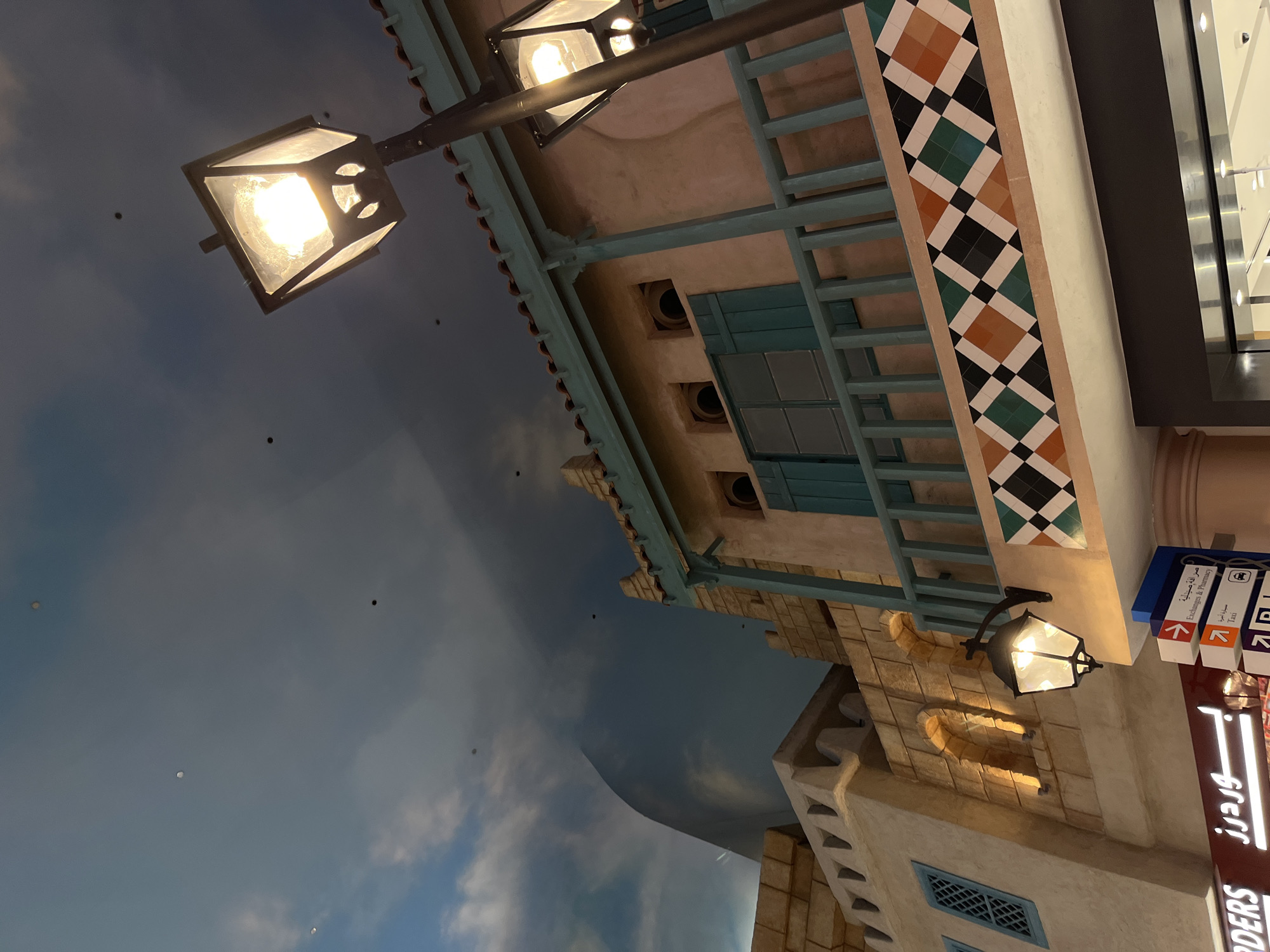 The image captures a section of a building with an outdoor facade, reminiscent of traditional Middle Eastern architecture, located in Dubai, United Arab Emirates. The building features beige stone walls, arched windows illuminated by interior lights, and a balcony with a light blue railing. Decorative tiles in a diamond pattern of orange, green, and black adorn the wall below the balcony. Two street lamps with glowing bulbs are visible, one attached to the building and another on a pole. In the background, a stylized sky with clouds is painted onto the ceiling, creating an illusion of an open sky. Signs in both Arabic and English are present at the bottom of the image, indicating directions to "Exchanges & Pharmacy" and "Taxi." The overall scene suggests an indoor recreation of an outdoor environment, likely within a shopping mall or entertainment complex in Dubai. There are no people visible in the image.