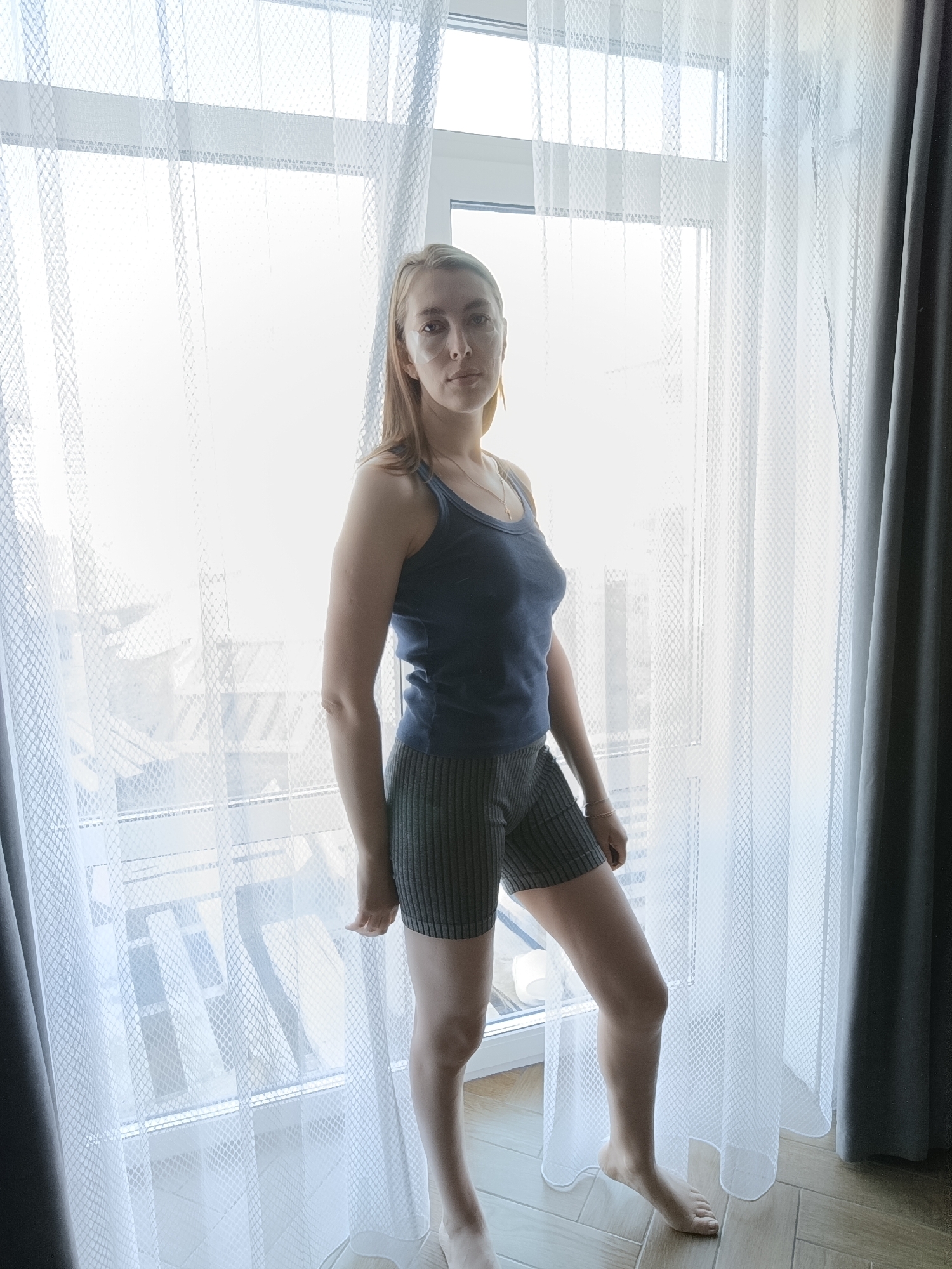 A woman with blonde hair stands indoors by a window with white, semi-transparent curtains. She wears a blue tank top and striped shorts. A small, white patch is visible on her left cheek. The floor appears to be tiled in a light brown, wood-grain pattern. Sunlight streams through the window. Dark gray curtains are to the right of the frame. Based on the available visual information, I cannot confirm if the photo was taken in the city of Chaikovsky, Russia, or any specific location.