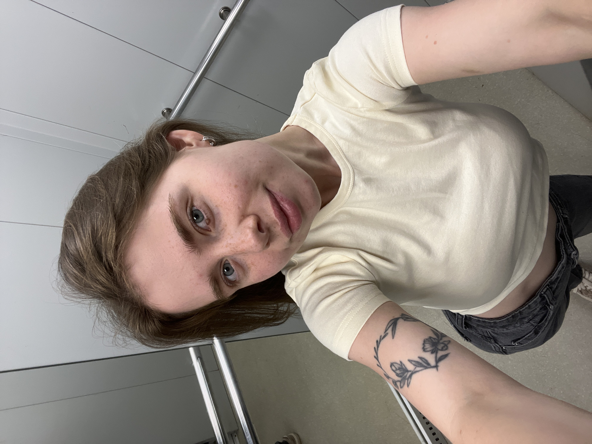 A young woman with light brown hair and blue eyes is taking a selfie in what appears to be an elevator. She has freckles on her face and is wearing a light yellow t-shirt and dark jeans. There is a tattoo of flowers on her left arm. The elevator walls are grey, and there are metal handrails on the sides. The floor is tiled. There is no specific indication in the image that it was taken in Chaikovsky, Russia.