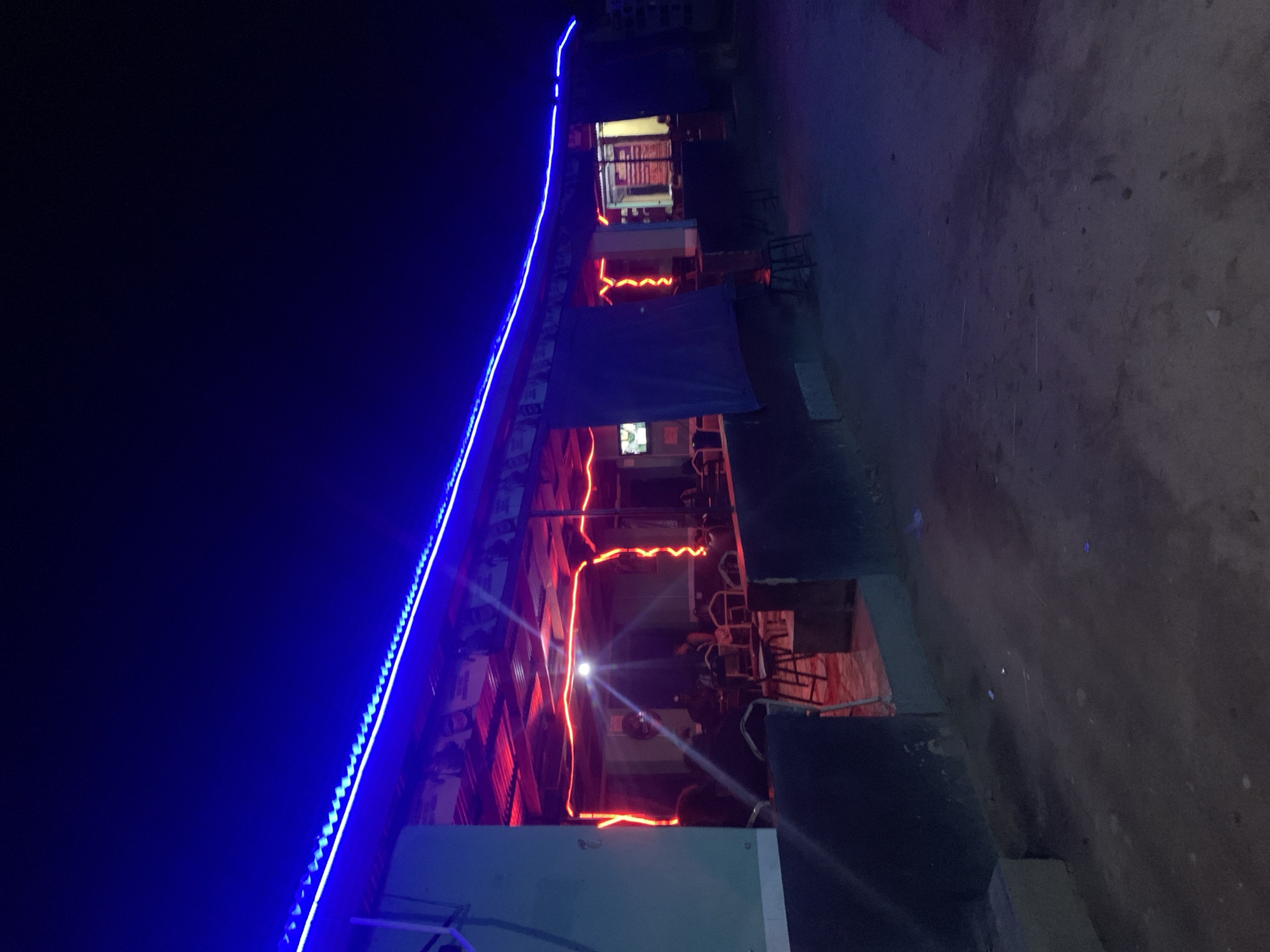 The image depicts a nighttime scene in what appears to be a commercial or entertainment area. There is a building with multiple levels illuminated by blue and red neon lights. The blue lights line the roof's edge, while the red lights seem to be outlining the interior of each level. There are tables and chairs visible within the building, suggesting a restaurant or cafe. The ground is dark and appears to be a dirt or gravel surface. The location is New Stadium, Nigeria.