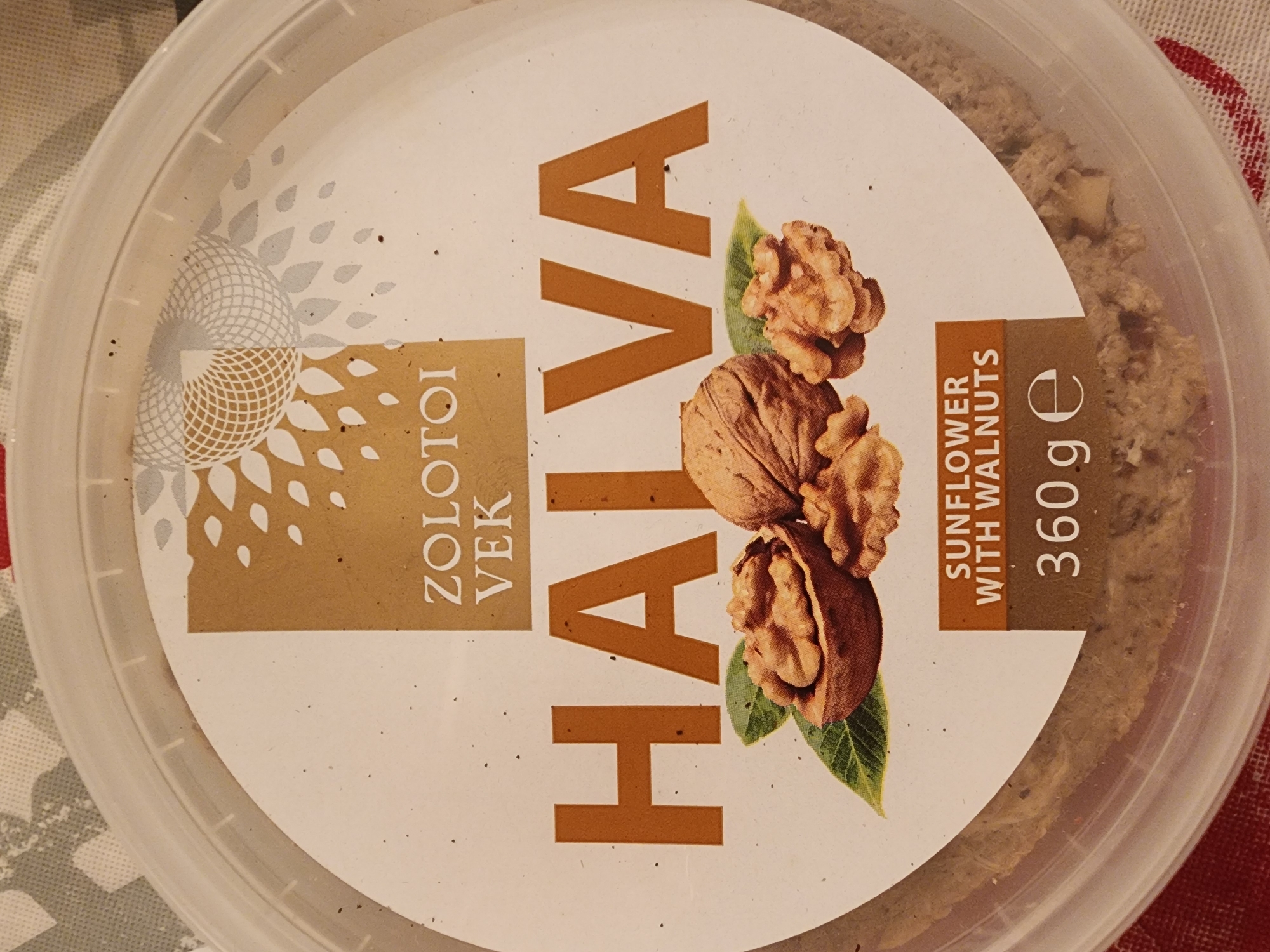 An overhead medium shot shows a round, clear plastic tub of halva on a red and white checkered surface. The lid of the tub has a label with a white background and a gold square in the upper left corner. Inside the gold square is a stylized white and gold sun-like graphic. Below the gold square, the text "ZOLOTΟΙ VEK" is printed in gold letters. The word "HALVA" is printed in large, bold, orange letters across the center of the label. Below "HALVA" are images of walnuts, one whole and two cracked open, with green leaves. Below the walnuts, a small orange rectangle contains the text "SUNFLOWER WITH WALNUTS" in white letters. Below this is the text "360 ge" in bold white letters. The halva inside the tub appears to be a light brown, crumbly substance with visible pieces of nuts.