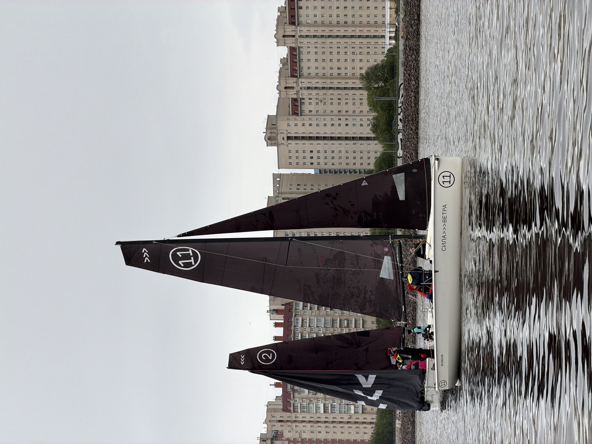The image shows a catamaran sailboat on the water in Saint Petersburg, Russia. Several tall residential buildings are visible on the shore in the background. The boat has three black sails, each with a white number: "2" on the bottom sail and "11" on the two larger sails. On the hull of the boat, it has the Russian text "СИЛА>>>ΒΕΤΡΑ", Russian text "RUS56126", and the number "11" within a circle. Multiple individuals can be seen sitting on the boat. The sky is overcast and grey. The water has reflective ripples.