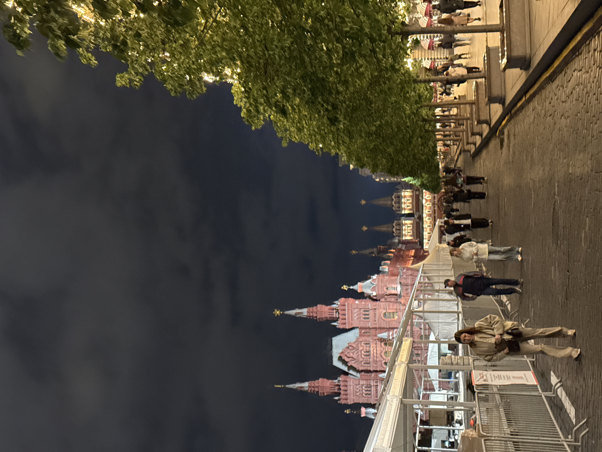 This nighttime scene in Moscow, Russia, depicts a street lined with trees adorned with lights. The distinctive spired architecture of the Kremlin is visible. Several individuals are walking along the cobblestone pavement, which is lined with metal barriers on one side. The sky is dark, suggesting a late hour. People are walking along the road, some with shopping bags.