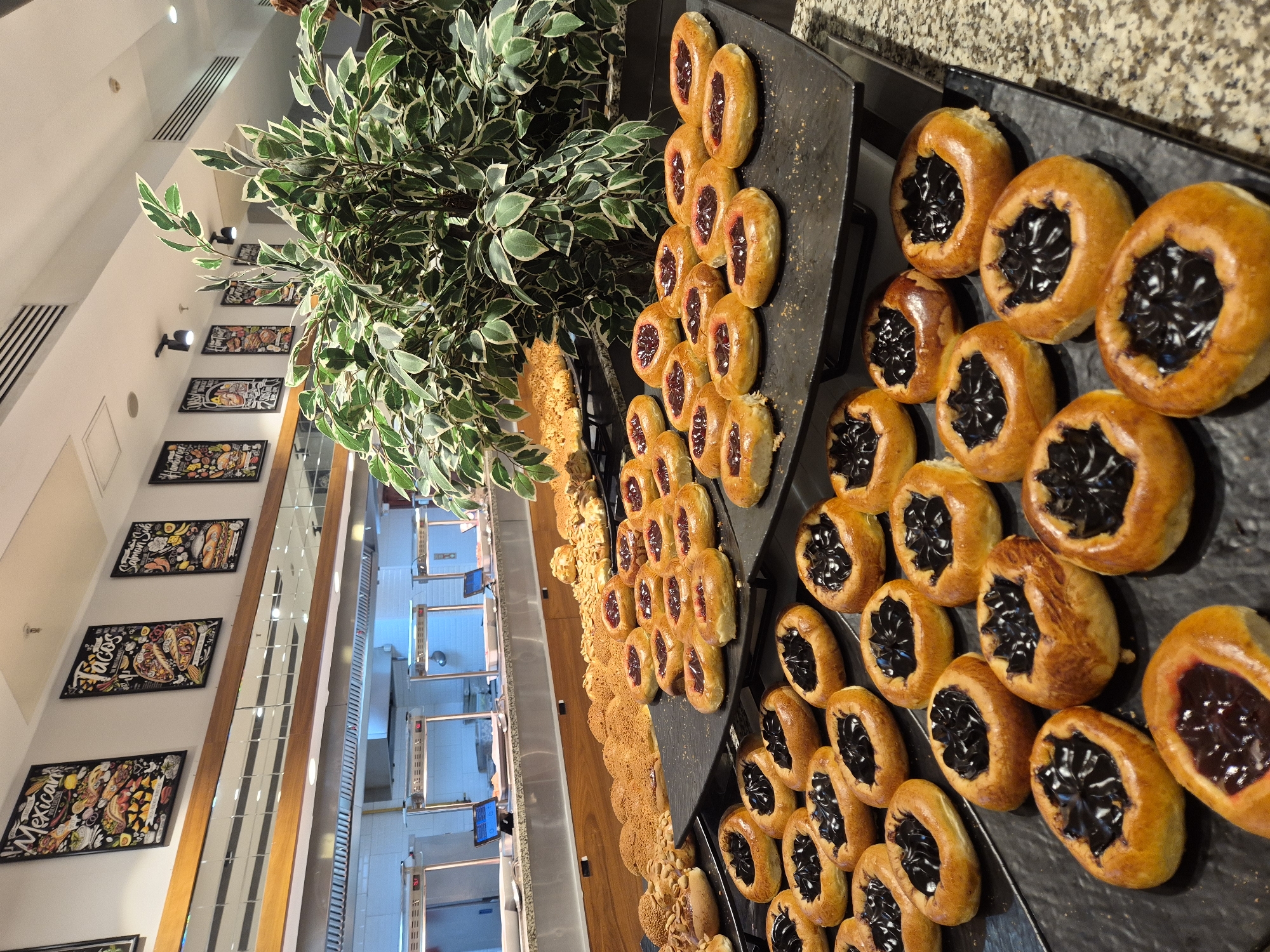 The image shows a close-up, high-angle view of a buffet display featuring a variety of baked goods. In the foreground and extending into the midground, a selection of pastries is arranged on dark platters. Some pastries are filled with a dark, jelly-like substance and have a golden-brown, slightly glossy crust. Other pastries appear to be filled with a red jam and have a similar golden-brown hue. Behind the pastries, a dense, leafy green plant with variegated leaves provides a backdrop.
Further in the background, a series of framed pictures hang on a white wall, adorned with illustrations of food items and text. One picture clearly displays the word "TACOS," another "MEXICAN," and a third "SALMON SUB." The overall setting appears to be an indoor food establishment, possibly a restaurant or cafe, with a modern aesthetic suggested by the white walls, recessed lighting, and mirrored panels in the distance which reflect the interior of the establishment. The baked goods are the primary focus, suggesting a scene of food service or display. There is no indication of people, time of day, weather, or emotions.