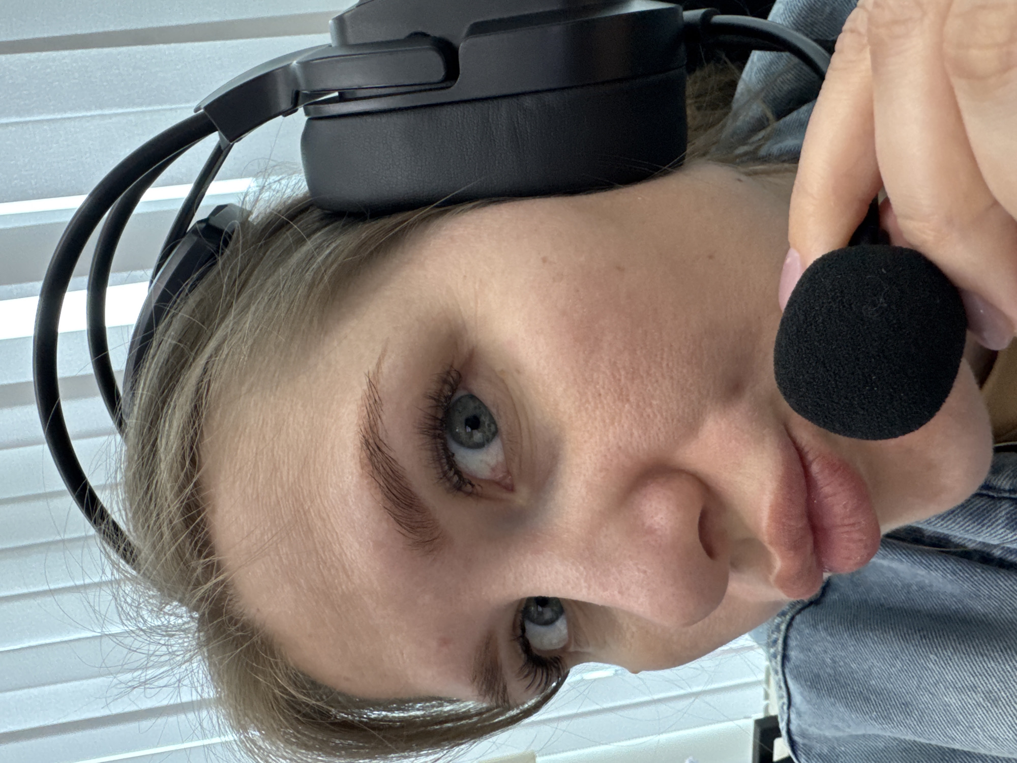 An indoor, portrait-style photograph captures a young adult woman wearing a black over-ear headset with a boom microphone. The image is presented sideways, with the woman's head tilted. She has light blue eyes, naturally defined eyebrows, and long eyelashes. Her light brown hair is visible around her temples and ears. Her lips are slightly pursed or pouting. Her right hand, adorned with light pink nail polish, holds the black foam-covered microphone close to her mouth. She is wearing a light blue denim jacket or shirt, visible on her right shoulder and arm. The background features white horizontal Venetian blinds, suggesting a window or room divider, with bright ambient light indicating daytime. No text is visible. The setting is in Perm, Russia.