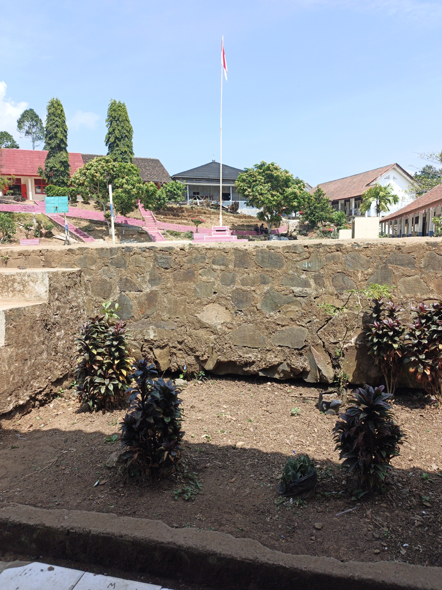 This is a medium-shot, eye-level, outdoor, daytime photograph taken in Tanjung Setia, Indonesia. The scene depicts a school courtyard with several buildings in the background under a clear blue sky with scattered clouds. In the foreground, a low, rough-hewn stone wall runs horizontally across the frame, separating the lower level from the upper level. In front of the wall, a patch of dry, brown soil is visible with three dark-leaved plants. To the left of the frame, a concrete structure with steps ascends.
In the mid-ground, the courtyard is tiered and features several buildings with red tiled roofs and white walls. The buildings appear to be school structures. A tall flagpole with the Indonesian flag is visible on the right side of the frame, and another similar flagpole is partially visible on the left. Pink-painted stairs and decorative elements are scattered throughout the mid-ground. Green trees are interspersed between the buildings and on the hillside.
The background is dominated by the sky. The image is a still shot, with no visible actions or interactions. The overall impression is of a typical school environment in a tropical location.