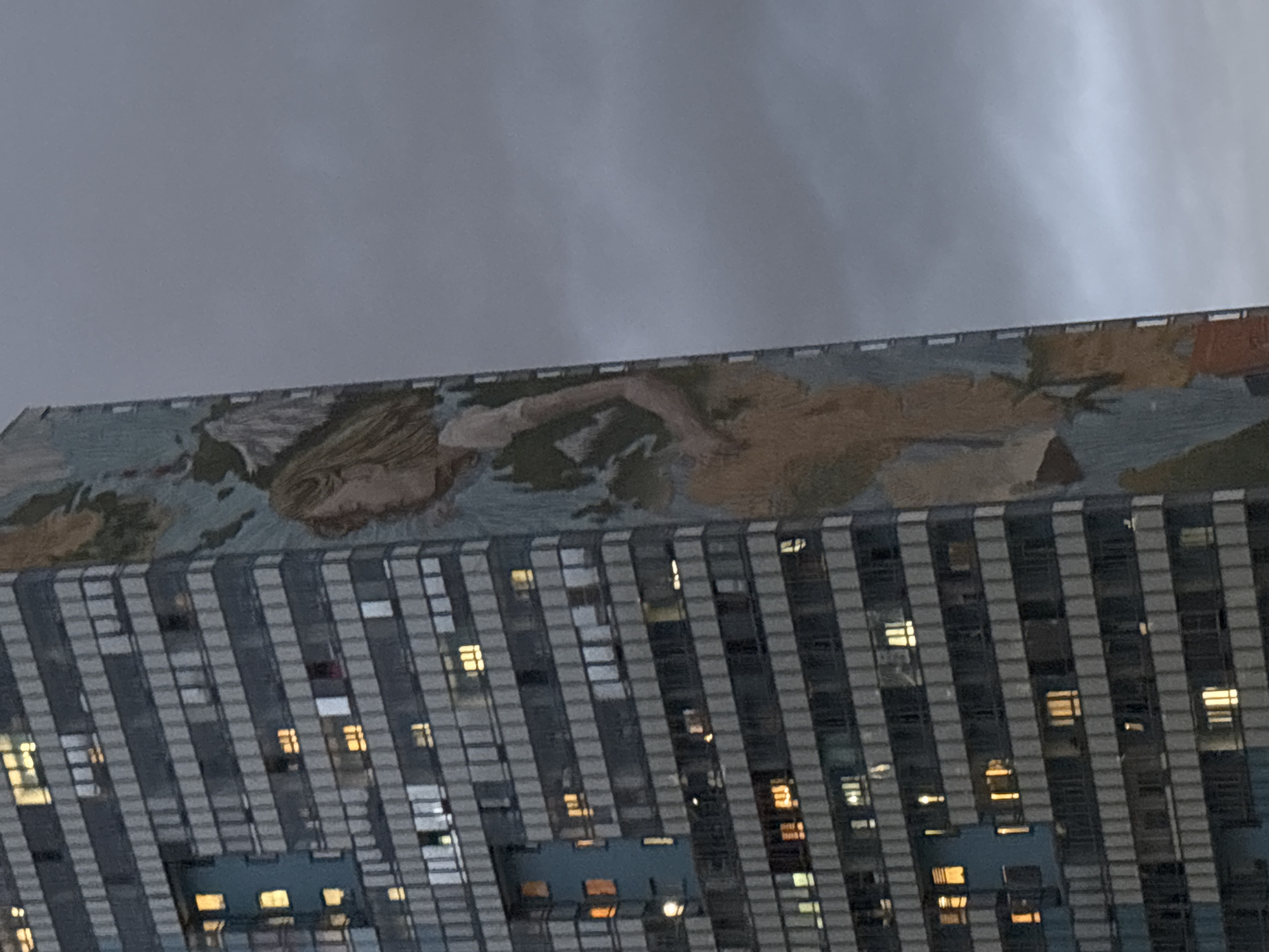 The image shows a building in Saint Petersburg, Russia. The building is tall and modern, with a grey and white striped facade and many windows, some of which are illuminated from within. At the top of the building, there is a large mural depicting a person with light hair lying on their side. The sky above the building is overcast.