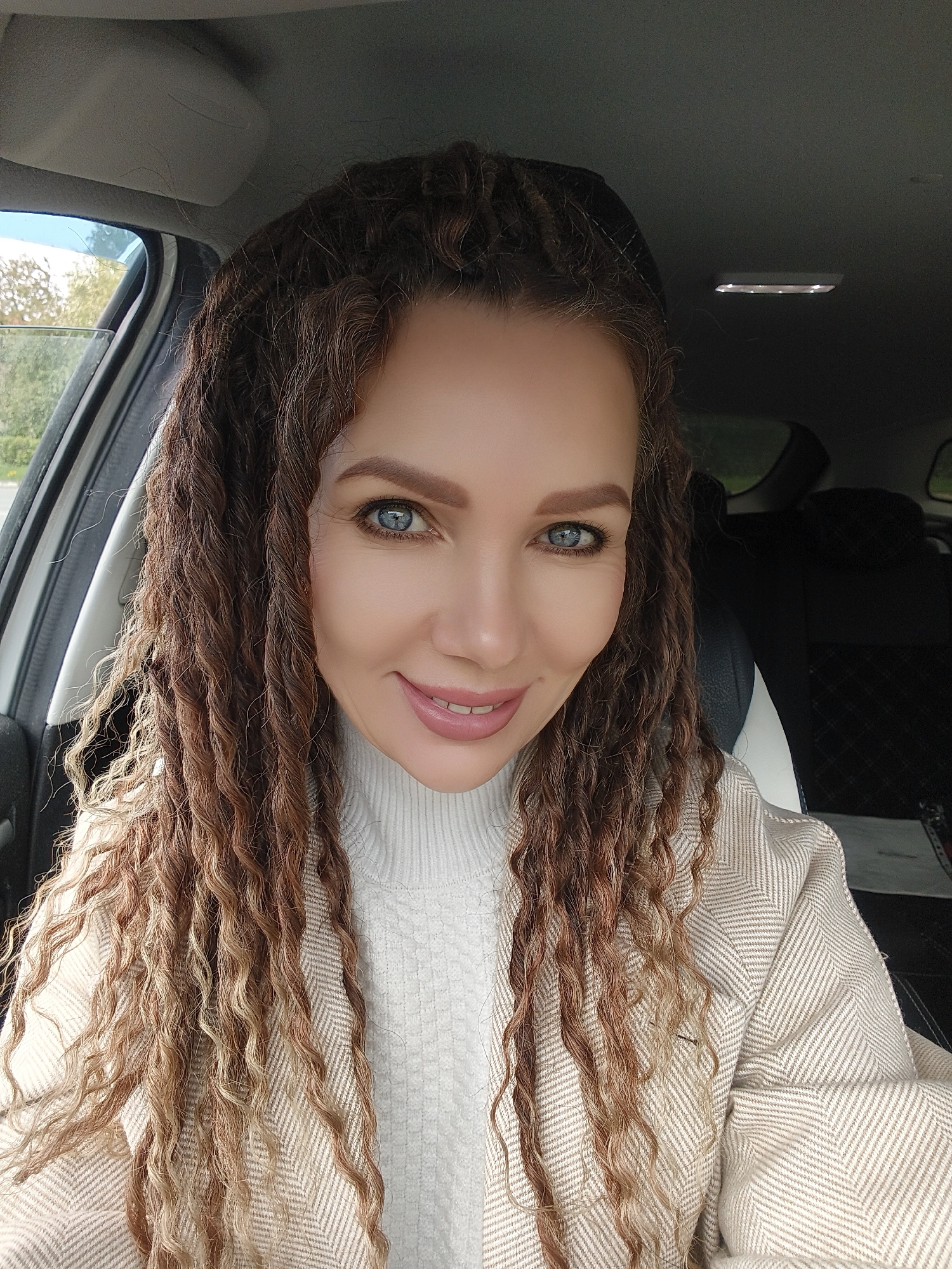 A woman is shown in a medium close-up shot from the chest up, likely inside a car in Unknown, Unknown. She has light skin, blue eyes, and brown eyebrows. Her hair is long and styled in a mix of dark brown braids and lighter curly strands. She is wearing a white turtleneck sweater and a beige herringbone pattern coat. She is smiling at the camera. The interior of the car is visible with white and black seats and a window in the background.